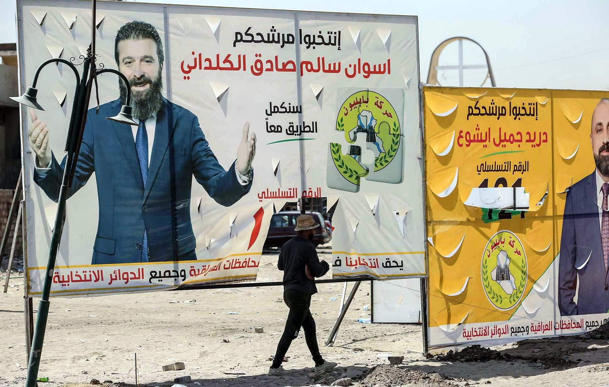 L'image montre un homme marchant devant de grandes affiches électorales dans un environnement urbain. Les affiches présentent des candidats politiques avec leurs noms écrits en arabe, ainsi que des logos et des slogans associés à leur campagne. L'arrière-plan semble désordonné, avec des structures inachevées ou des éléments typiques d'une ville. L'homme semble porter une charge sur sa tête, ajoutant une dimension de vie quotidienne à la scène.