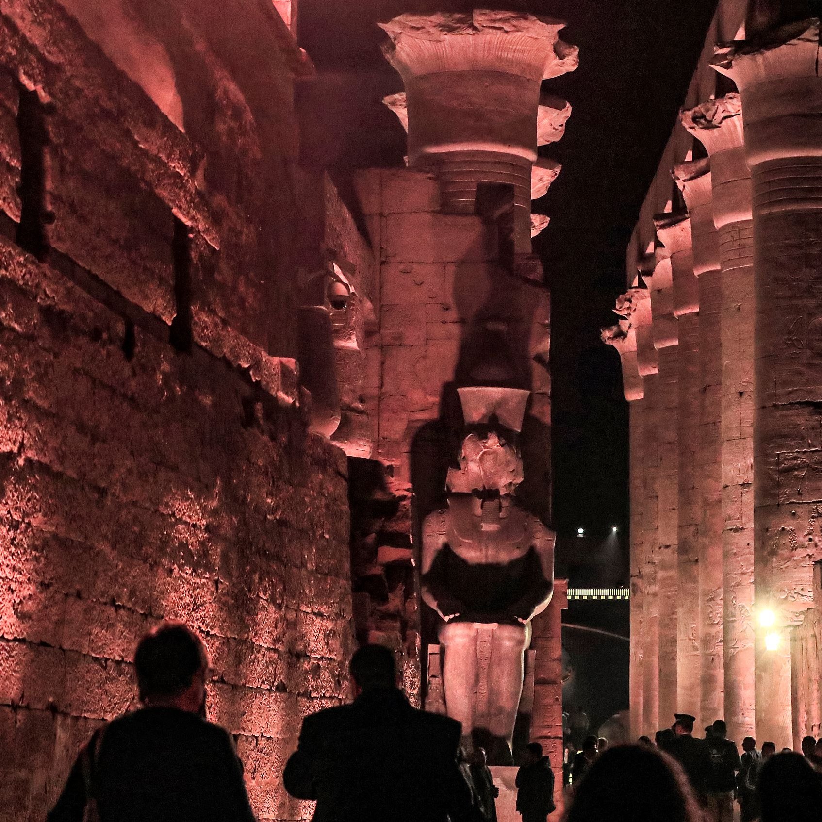 The image depicts a nighttime scene at a historic location, likely a temple or ancient site, characterized by large stone structures. Towering columns line the pathway, illuminated by soft red and amber lighting, which casts dramatic shadows on the walls. Two imposing statues, possibly representing pharaohs or deities, stand prominently at the end of the corridor. Visitors can be seen walking through the area, taking in the majestic architecture and ambiance of this ancient site. The atmosphere conveys a sense of history and reverence.