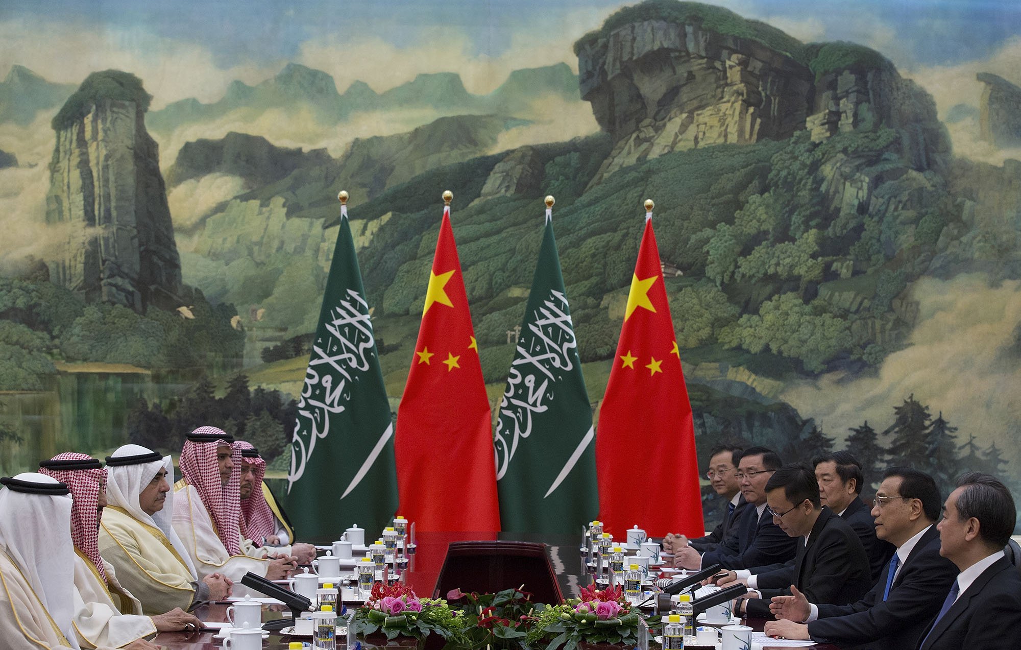 در این تصویر، یک نشست رسمی بین مقامات سعودی و چینی در حال برگزاری است. دو طرف میز مذاکره نشسته‌اند و پرچم‌های عربستان سعودی و چین به وضوح در پس‌زمینه دیده می‌شود. فضای جلسه به گونه‌ای طراحی شده که یک پس‌زمینه طبیعی با کوه‌ها و درختان وجود دارد. هر طرف میز با تعداد زیادی فنجان و ظروف غذا چیده شده است و به نظر می‌رسد که محتوای بحث و تبادل نظر جدی و مهم باشد.