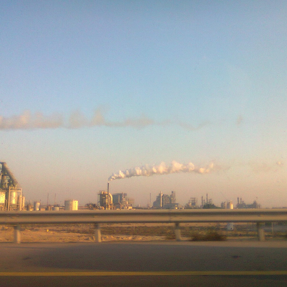 The image depicts an industrial landscape with several factories. In the foreground, there is a highway barrier, suggesting the viewer is looking from a moving vehicle. The factories emit smoke and steam into the atmosphere, creating white clouds against a clear blue sky. The overall scene conveys an industrial environment, typical of urban or semi-urban areas where manufacturing activities take place.
