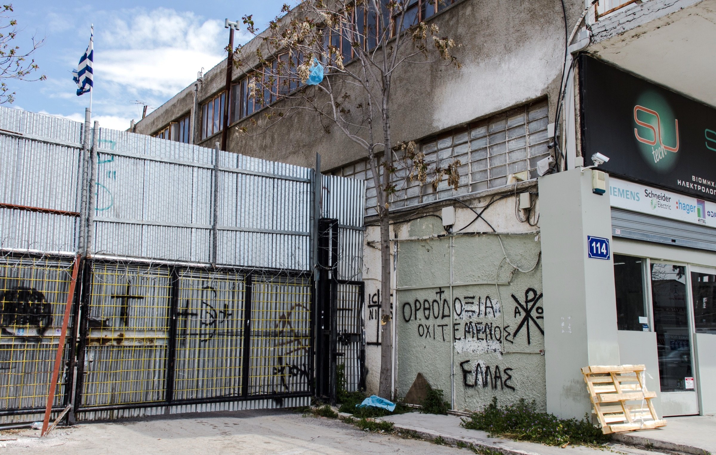 L'image montre un environnement urbain avec un bâtiment en arrière-plan et un mur métallique en façade, où l'on peut voir des graffitis. Un drapeau grec est visible, ainsi qu'un espace de stationnement ou de stockage à côté d'une entrée. Le sol semble en béton, et quelques déchets sont éparpillés sur le sol. Les graffitis sur le mur comprennent des inscriptions en grec, ce qui suggère un contexte local ou culturel. L'atmosphère paraît un peu désordonnée, typique des zones industrielles ou abandonnées.