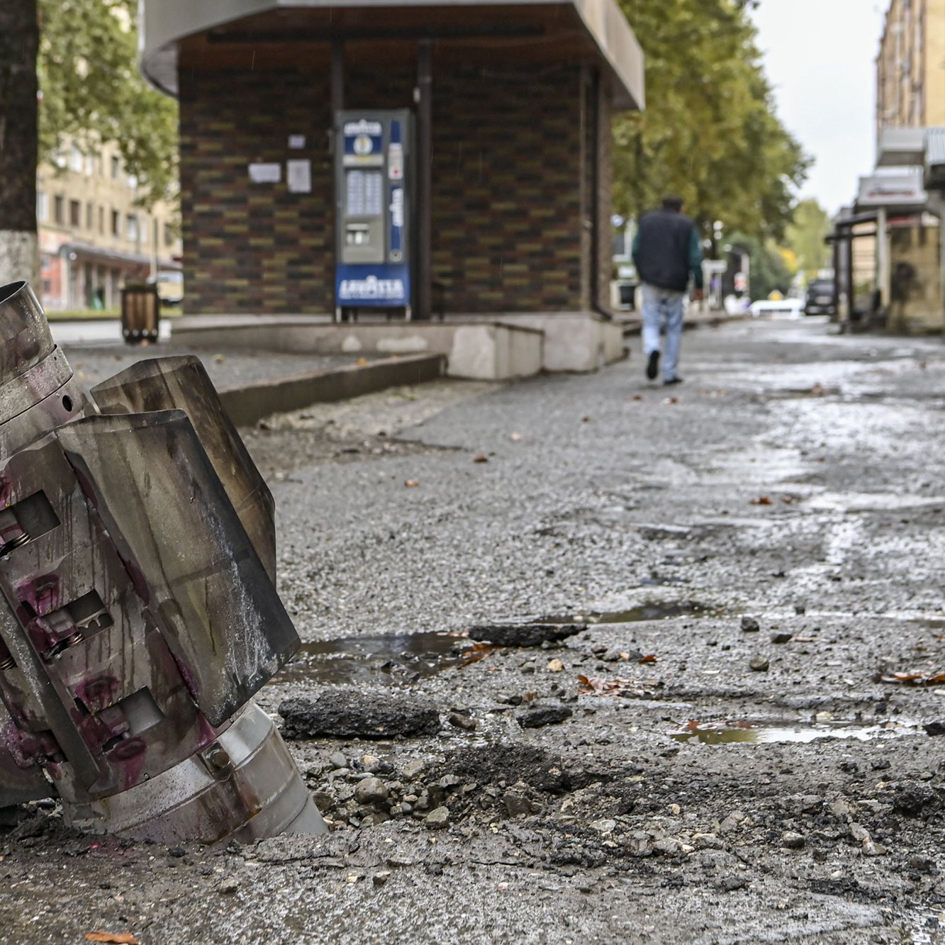L'image montre un paysage urbain marqué par la destruction. Au premier plan, on voit un obus ou un engin explosif détérioré, partiellement enfoui dans le sol. Le bitume est endommagé autour de l'obus, laissant des fissures et des débris. En arrière-plan, un homme marche sur le trottoir, près d'un bâtiment qui semble intact. L'atmosphère est sombre et témoigne des conséquences d'un conflit ou d'un événement violent. Les arbres sur le bord de la route ajoutent une touche de verdure, mais le contexte reste très préoccupant.