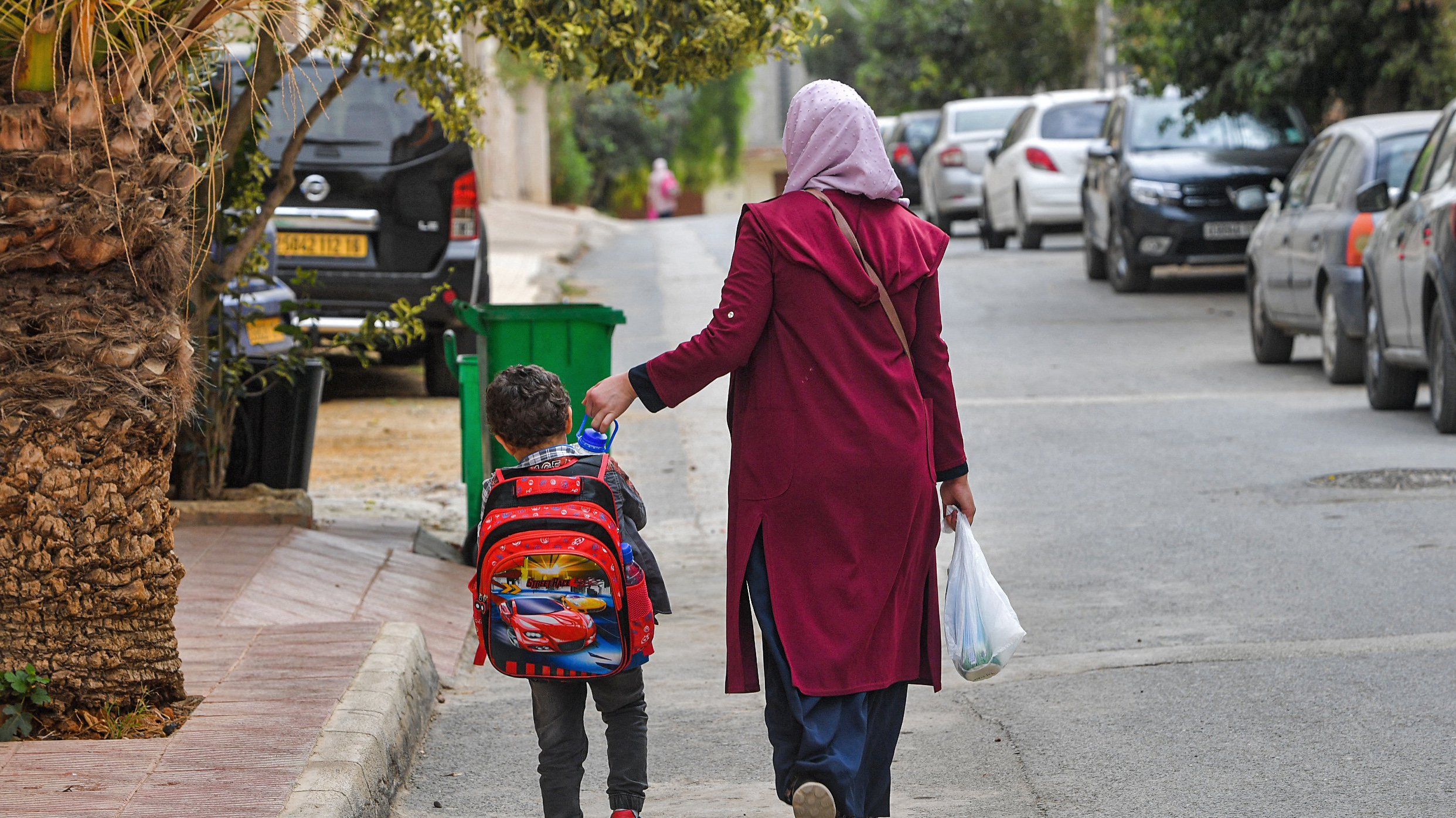 L'image montre une femme portant un foulard, vêtue d'un long manteau, qui marche dans une rue résidentielle avec un jeune garçon. Le garçon porte un sac à dos coloré, probablement inspiré d'un personnage de dessin animé. Ils semblent se diriger vers une destination, et la femme tient également un sac en plastique. En arrière-plan, on peut voir des voitures garées des deux côtés de la rue et de la verdure autour. L'atmosphère semble calme et quotidienne.