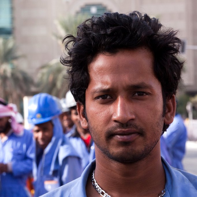 L'image montre un groupe de travailleurs, avec un homme au premier plan qui a les cheveux frisés et un regard intense. Il porte une tenue de travail bleue et une chaîne autour du cou. En arrière-plan, d'autres travailleurs en uniformes et casques bleus sont visibles, discutant entre eux. L'environnement semble urbain, avec des bâtiments modernes.