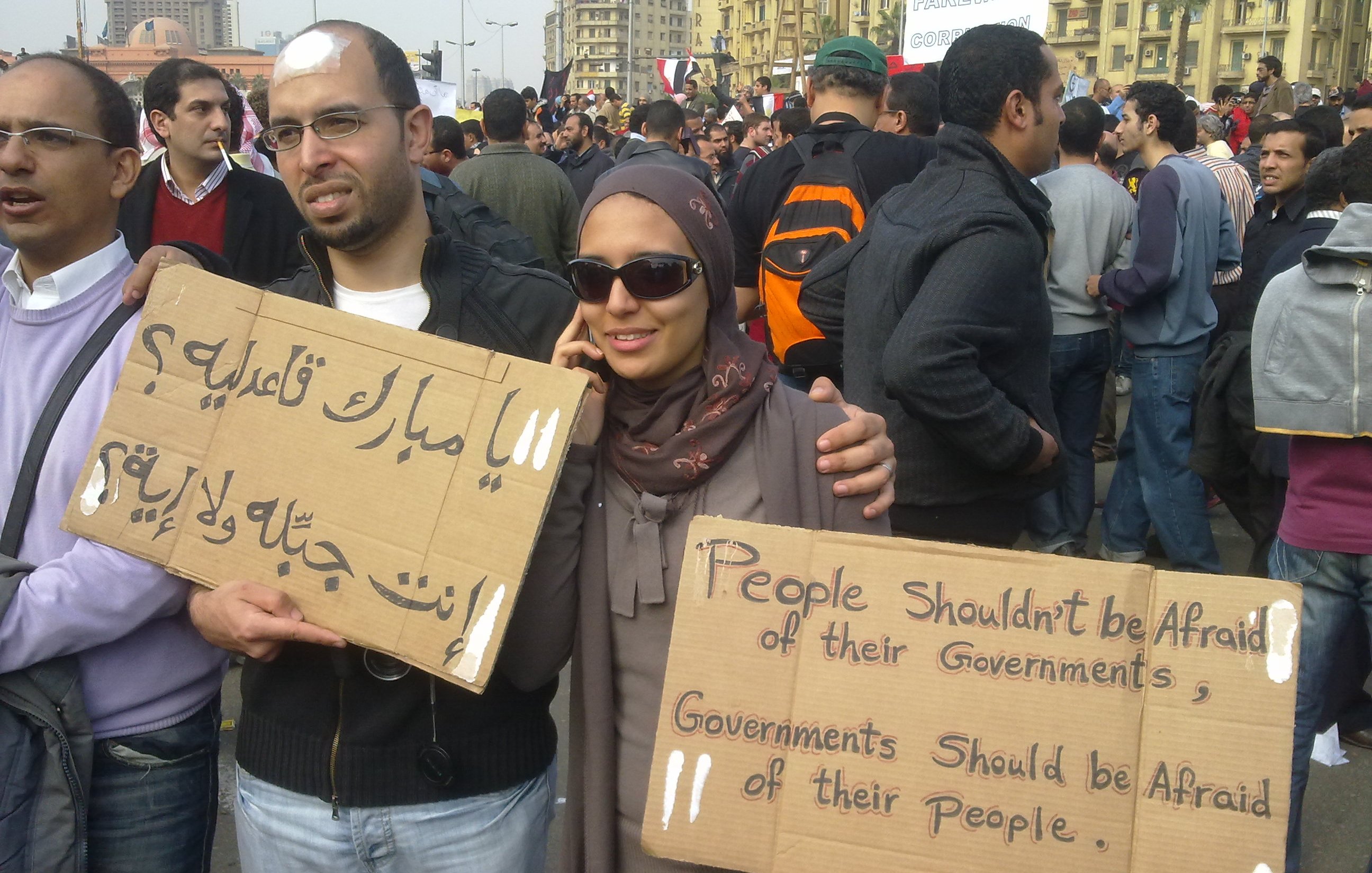 L'immagine mostra un gruppo di persone in una manifestazione. Due individui in primo piano tengono in mano dei cartelli. Uno dei cartelli è scritto in arabo e l'altro in inglese. Il messaggio in inglese dice "Le persone non dovrebbero avere paura dei loro governi. I governi dovrebbero avere paura del loro popolo." I partecipanti sembrano impegnati e determinati, con un'ampia folla di persone sullo sfondo. La scena evoca un senso di protesta e richiesta di cambiamento sociale.