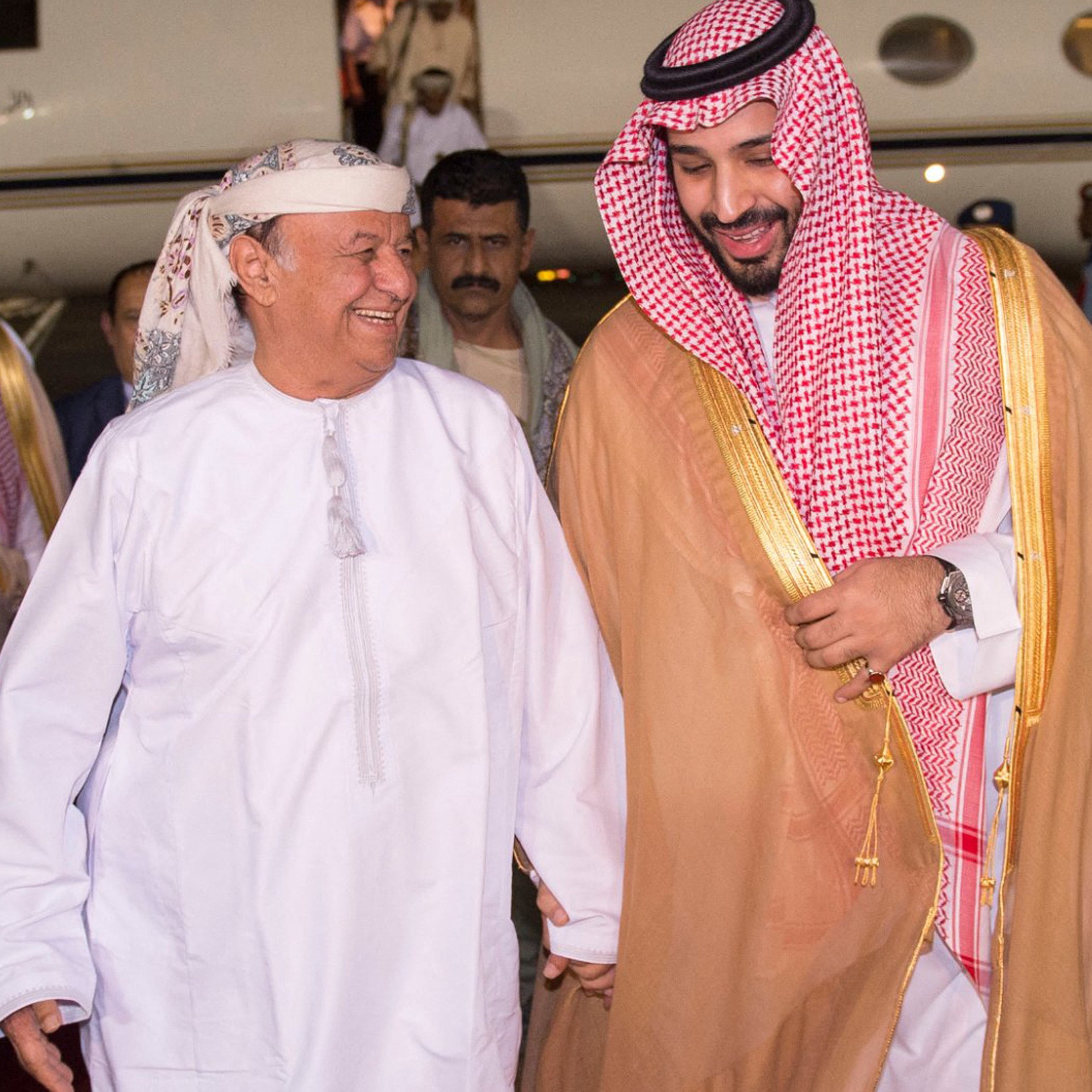 The image shows two men smiling and engaged in a friendly conversation as they walk towards a private jet. One man is dressed in traditional Arab attire, including a white thobe and a headscarf, while the other is wearing a similar traditional outfit, accented with a golden cloak and a checkered headscarf. They appear to be happy and are holding hands, suggesting a warm greeting. In the background, there are several individuals, likely staff or security, standing by the jet. The setting seems to be an airport or a private aviation terminal.