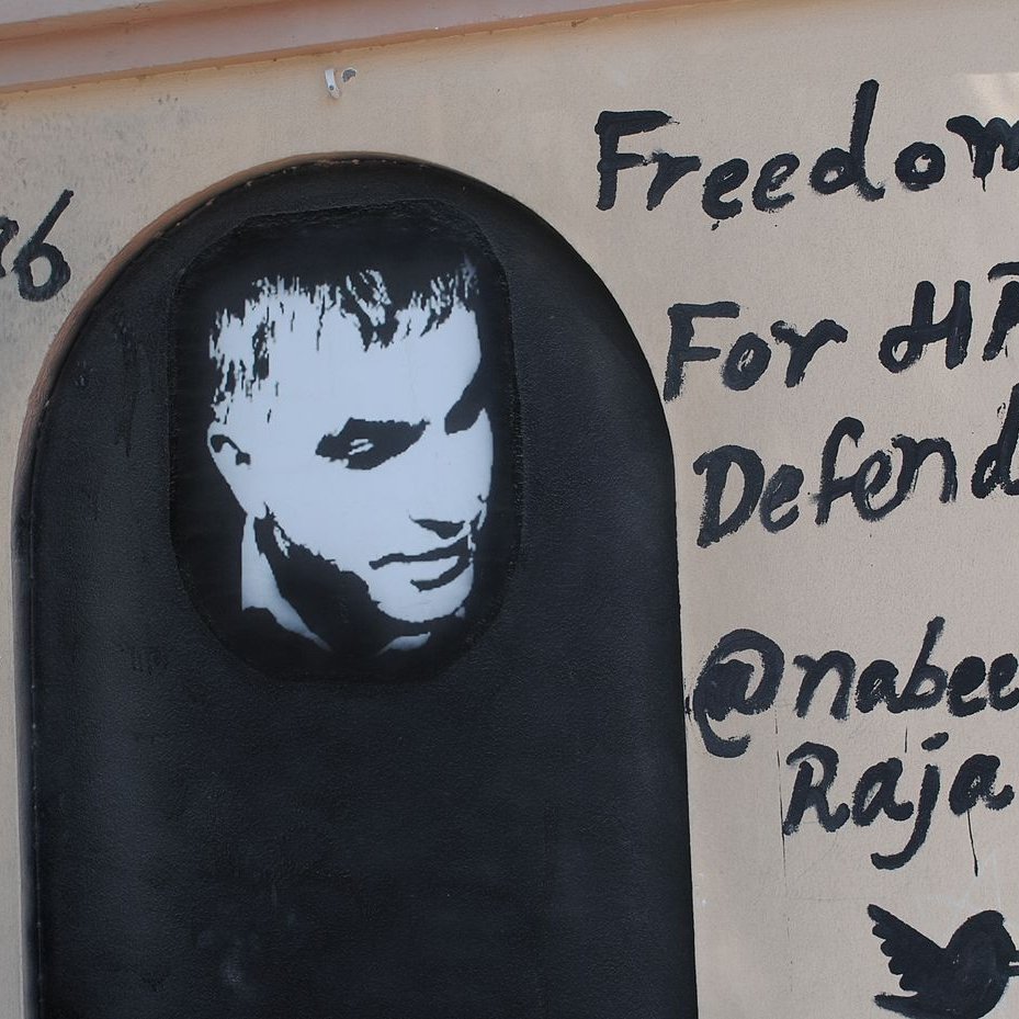 L'image représente un graffiti sur un mur. On y voit un portrait en noir et blanc d'un homme, accompagné d'un texte écrit en anglais. Le message indique "Freedom For HR Defender" (Liberté pour le défenseur des droits de l'homme) et mentionne un nom d'utilisateur Twitter "@nabeelrajab". Le style artistique est simple, avec un fond sombre et des lettres noires qui ressortent sur la surface claire du mur. Cela semble être une œuvre engagée, soulignant le soutien à la défense des droits humains.