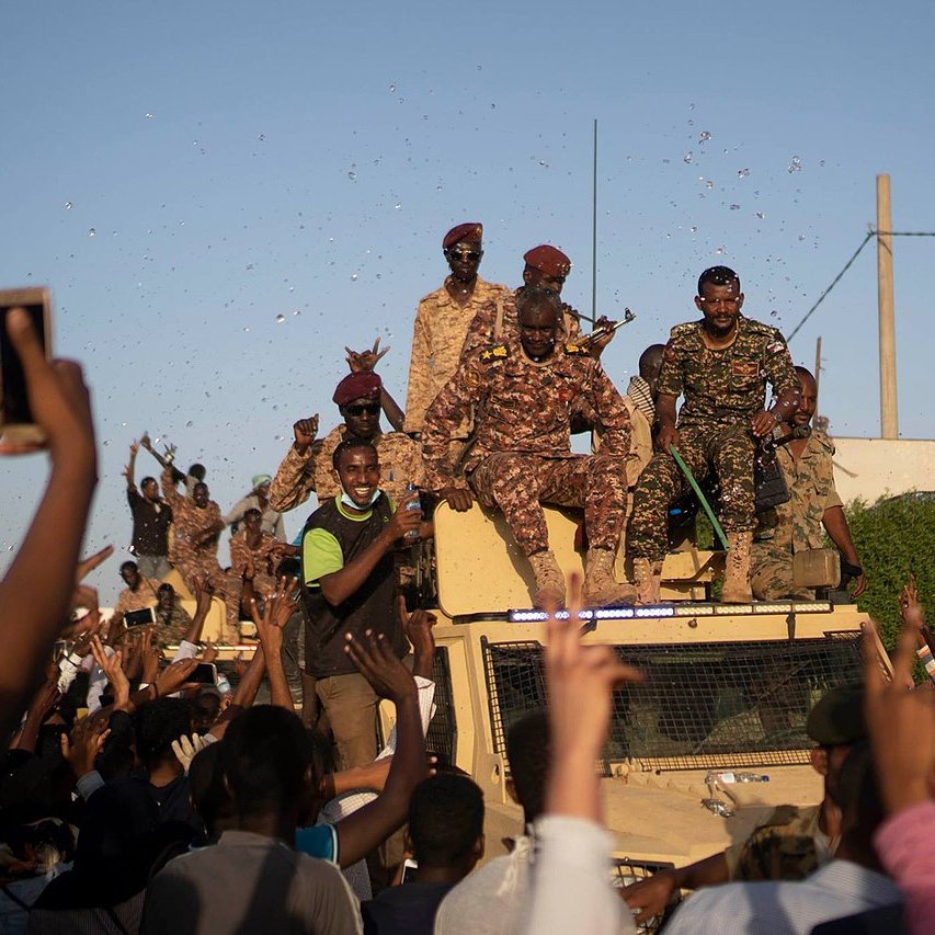 الصورة تظهر مجموعة من الجنود يقفون على متن مركبة عسكرية، بينما يبدو أن حشودًا من الناس تتجمع حولهم. يبدو الجو احتفاليًا حيث يرفع بعض الأشخاص أيديهم في الهواء، ويبدو أنهم يعبرون عن الفرح أو التأييد. السماء زرقاء وظهرت بعض الأعلام والاحتفالات في الخلفية.