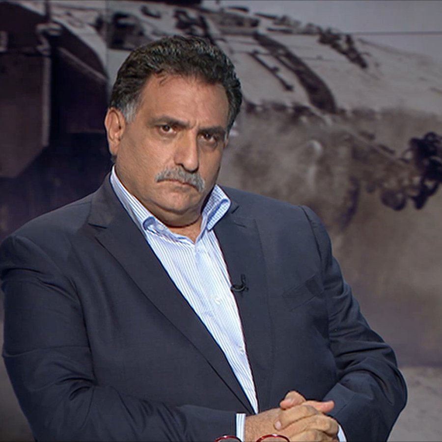 الصورة تظهر رجلًا يجلس بتعابير وجه جدية. يرتدي بدلة داكنة وقميص أبيض، ويداه متشابكتان أمامه. في الخلفية، يظهر مشهد غامض لمركبة عسكرية أو صورة حربية، مما يضيف جوًا من التوتر والدرامية للصورة. يبدو أن الرجل يتأمل أو يفكر بعمق.