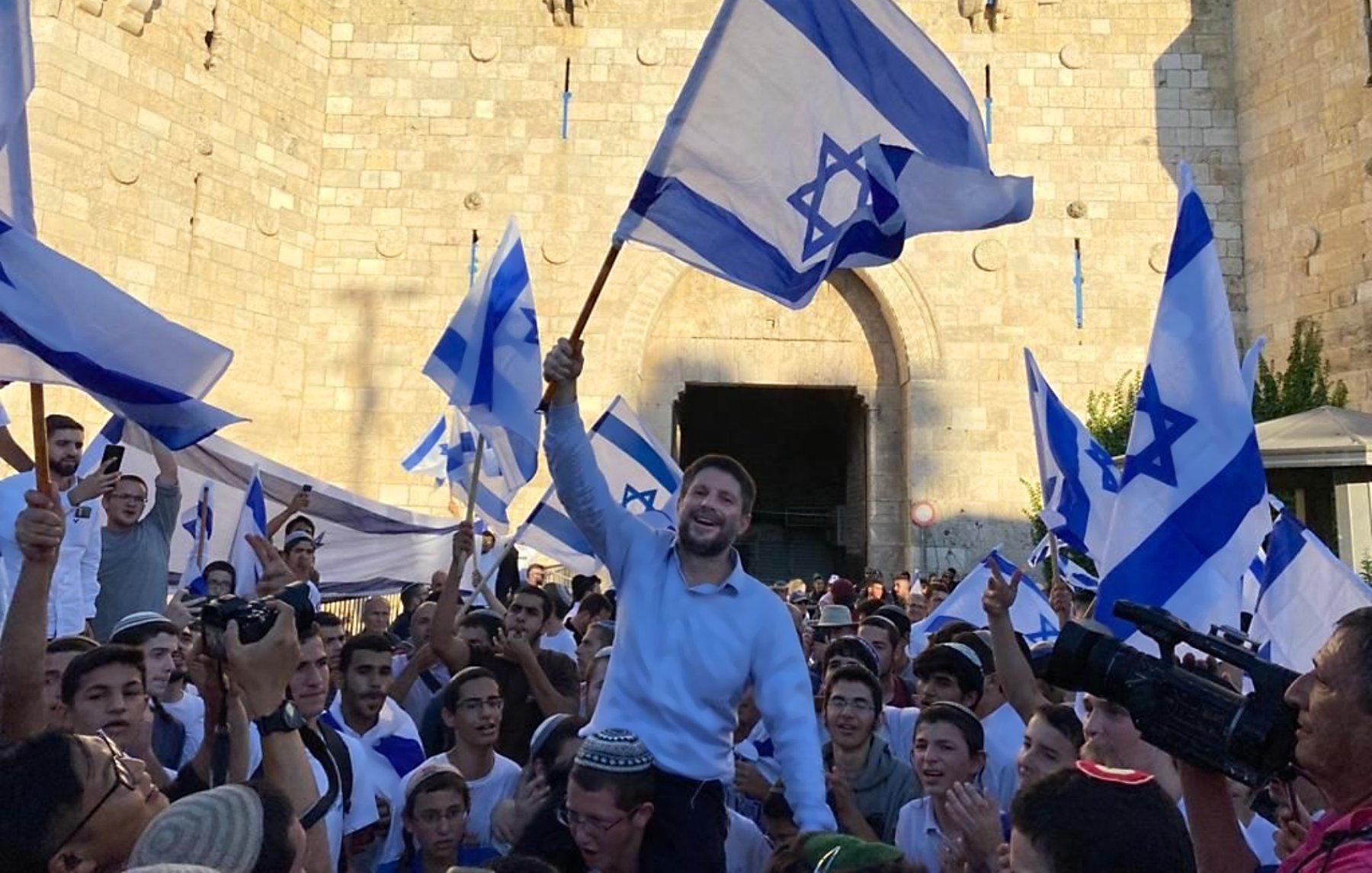 L'image montre une foule rassemblée devant une grande porte en pierre, probablement située dans une ville historique. Les participants tiennent des drapeaux bleu et blanc, représentant l'État d'Israël. Un homme se tient au centre, soulevant un drapeau avec un sourire, tandis que d'autres l'entourent, festoyant. L'atmosphère semble festive et joyeuse, suggérant une célébration ou un événement important.
