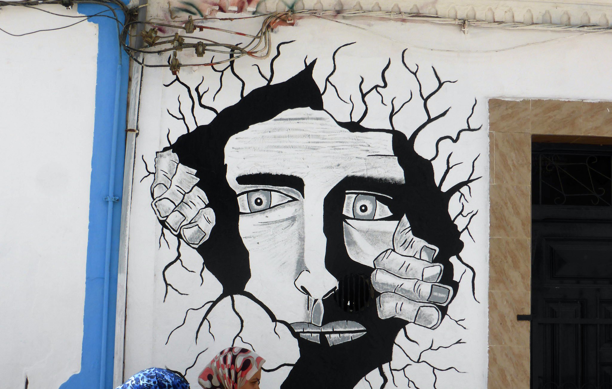 تظهر في الصورة جداريات فنية باللونين الأبيض والأسود، حيث يبرز وجه إنسان يبدو كأنه يحاول الخروج من جدار. يحيط بالوجه شقوق في الجدار، مما يضفي شعورًا بالديناميكية والحركة. في الجزء السفلي من الصورة، يوجد امرأتان تسيران بجوار الجدارية، مما يعكس تفاعل الحياة اليومية مع الفن المعاصر. الجدارية تحمل توقيع الفنان "توفيق" بالإضافة إلى حسابه على إنستغرام.