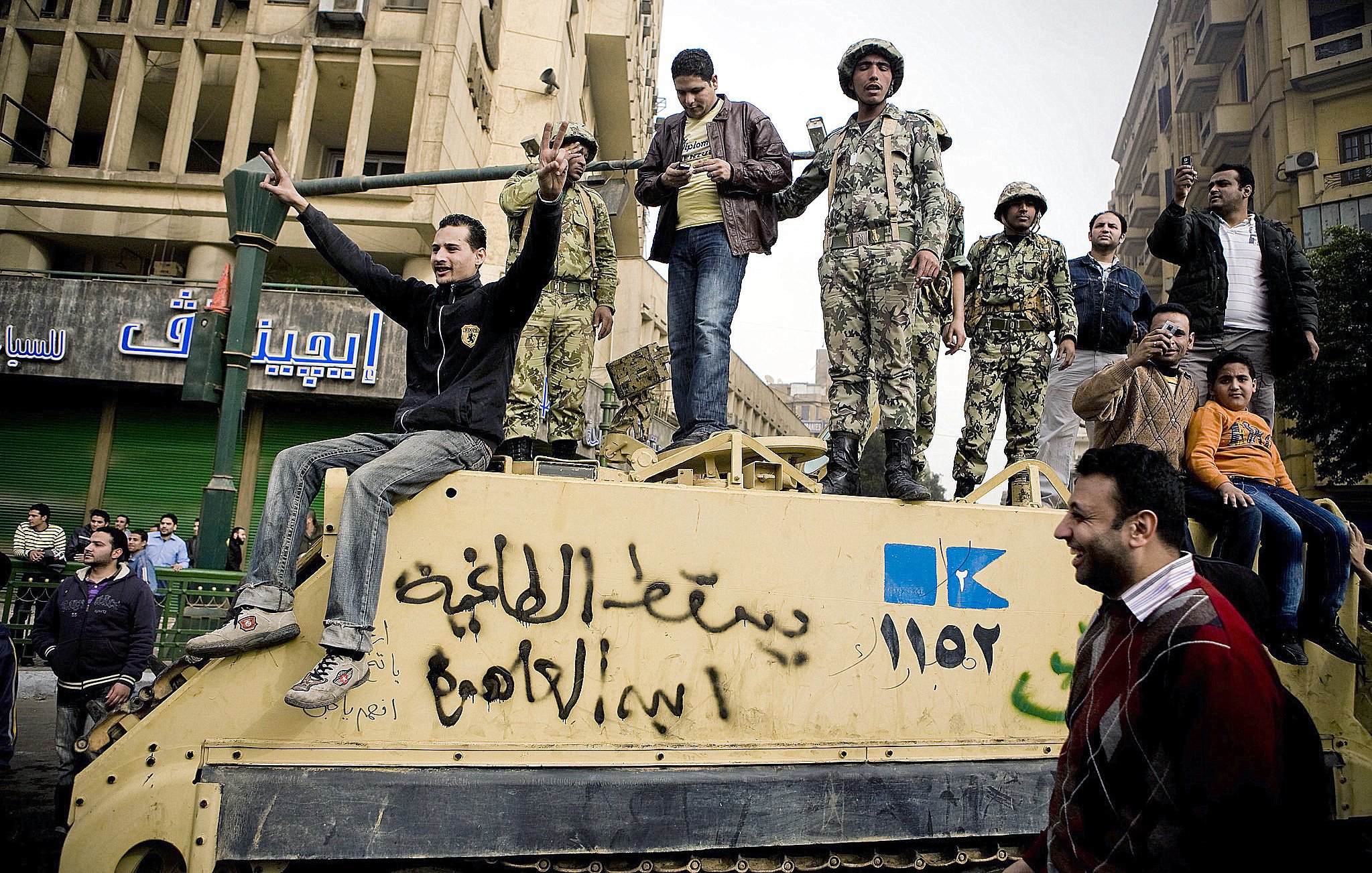 L'image montre un groupe de personnes rassemblées autour d'un véhicule militaire dans une ville. On peut voir des soldats en uniforme sur le véhicule, tandis que d'autres personnes, probablement des civils, interagissent avec eux. L'atmosphère semble tendue, mais aussi pleine d'énergie, comme si une sorte de manifestation ou de rassemblement avait lieu. L'arrière-plan montre des bâtiments urbains et des enseignes en arabe, ce qui suggère que la scène se déroule dans un pays du Moyen-Orient.