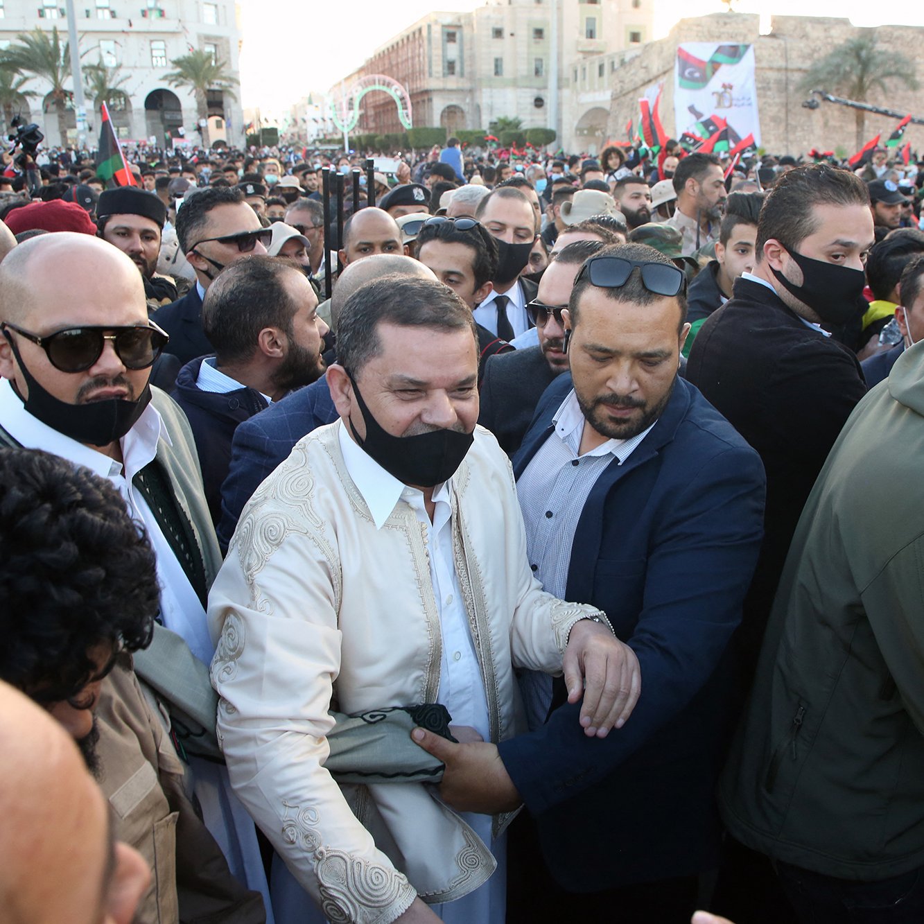 L'image montre une foule dense, avec un groupe de personnes rassemblées lors d'un événement. Au centre, un homme en tenue traditionnelle est entouré de plusieurs personnes portant des masques. L'ambiance semble être celle d'un rassemblement ou d'une célébration. Les participants affichent des expressions d'enthousiasme, certains tenant des bannières ou des drapeaux. En arrière-plan, des bâtiments et de la verdure sont visibles, ce qui indique une atmosphère festive.