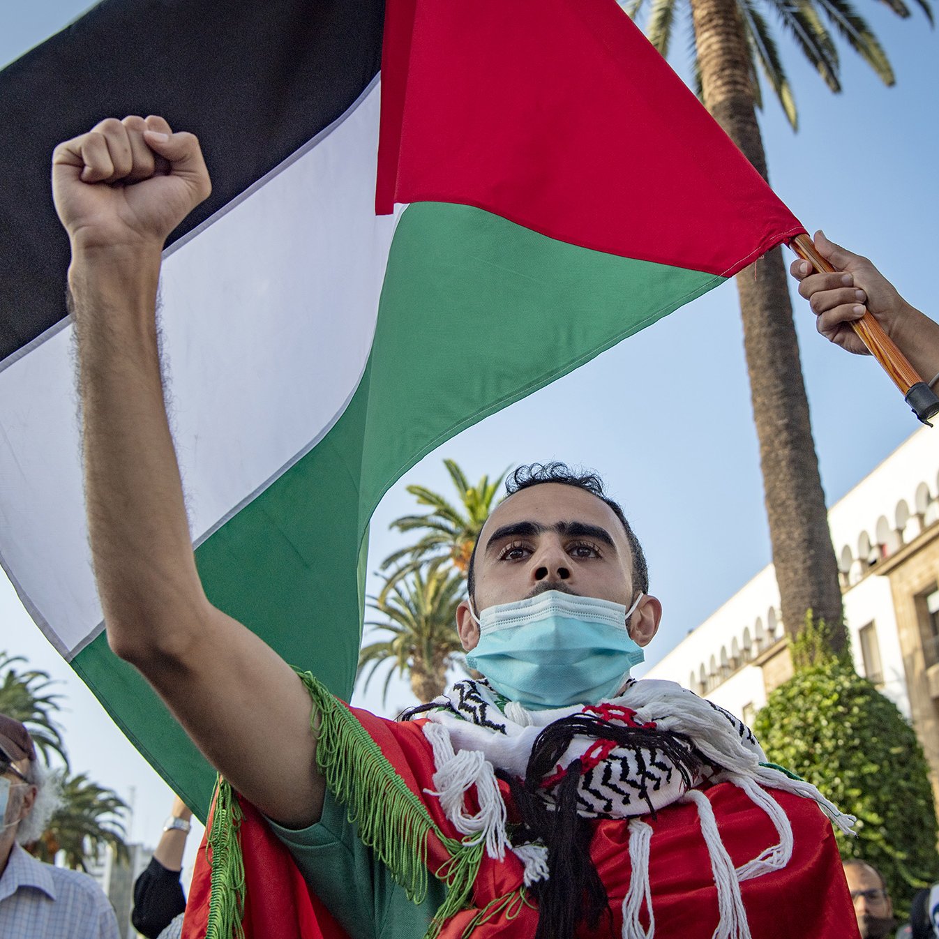 L'image montre une manifestation où un homme brandit fièrement un drapeau palestinien. Il porte un masque et est entouré de palmiers, ce qui indique un environnement ensoleillé. Son expression montre une détermination forte, et il semble engagé dans une cause importante. D'autres personnes sont présentes dans le fond, ajoutant au contexte collectif de la protestation.