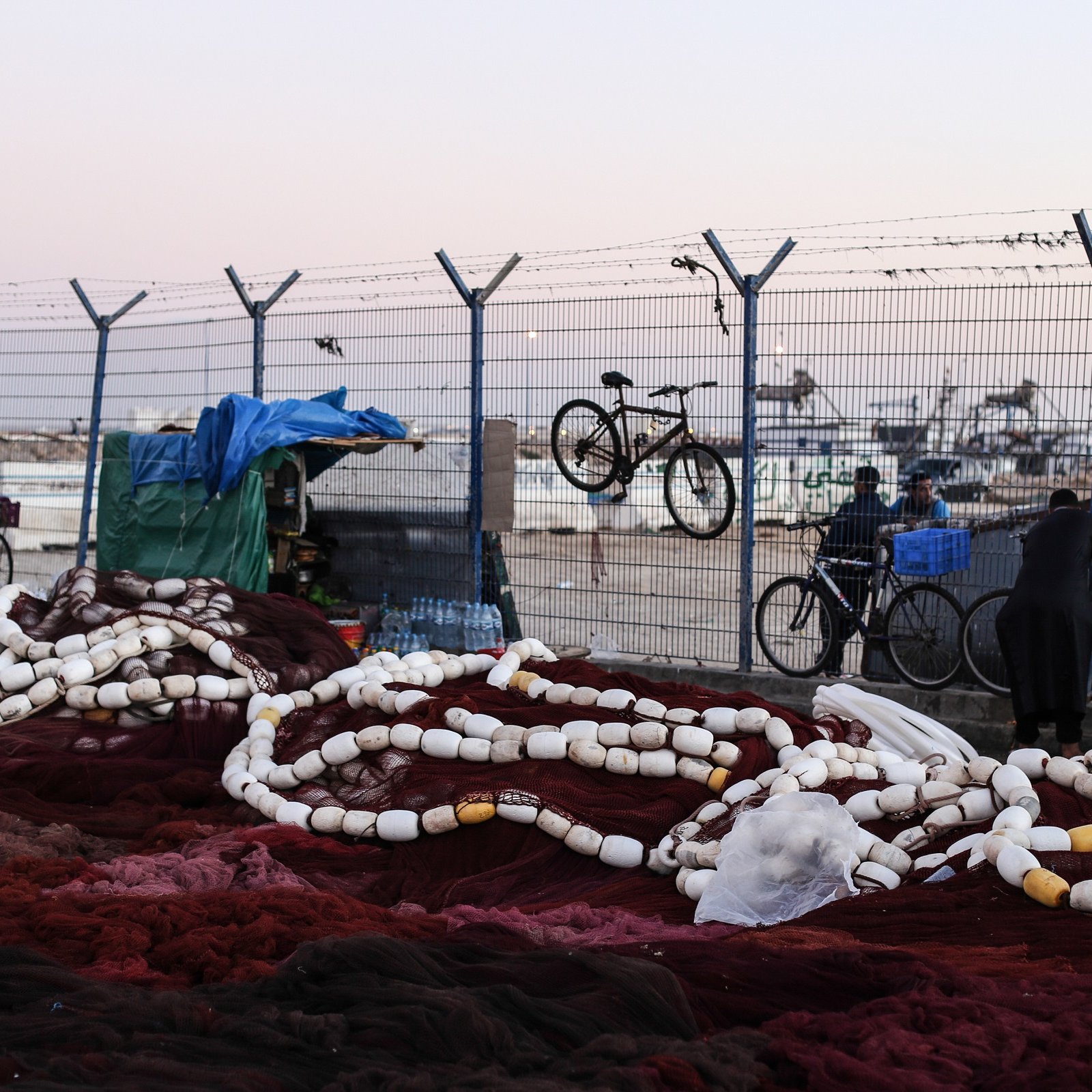 L'image montre un environnement portuaire où se trouvent des filets de pêche. De grandes cordes blanches, probablement des bouées, sont éparpillées sur le sol, mêlées à des filets de couleur rouge foncé. En arrière-plan, on aperçoit une clôture barbelée qui délimite l'espace, ainsi que quelques bicyclettes accrochées. On peut aussi voir des personnes se déplacer, l'une d'elles étant vêtue d'une tenue traditionnelle. Le ciel est légèrement teinté, indiquant peut-être le début de la soirée. L'atmosphère semble calme, mais il y a une impression d'activité liée à la pêche.