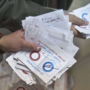 L'image montre des mains tenant des bulletins de vote. On peut voir des bulletins blancs portant des cercles rouges et bleus, ainsi que des inscriptions en arabe. Il semble s'agir d'une scène de vote, probablement lors d'une élection. Les bulletins sont rassemblés, suggérant un processus électoral en cours.