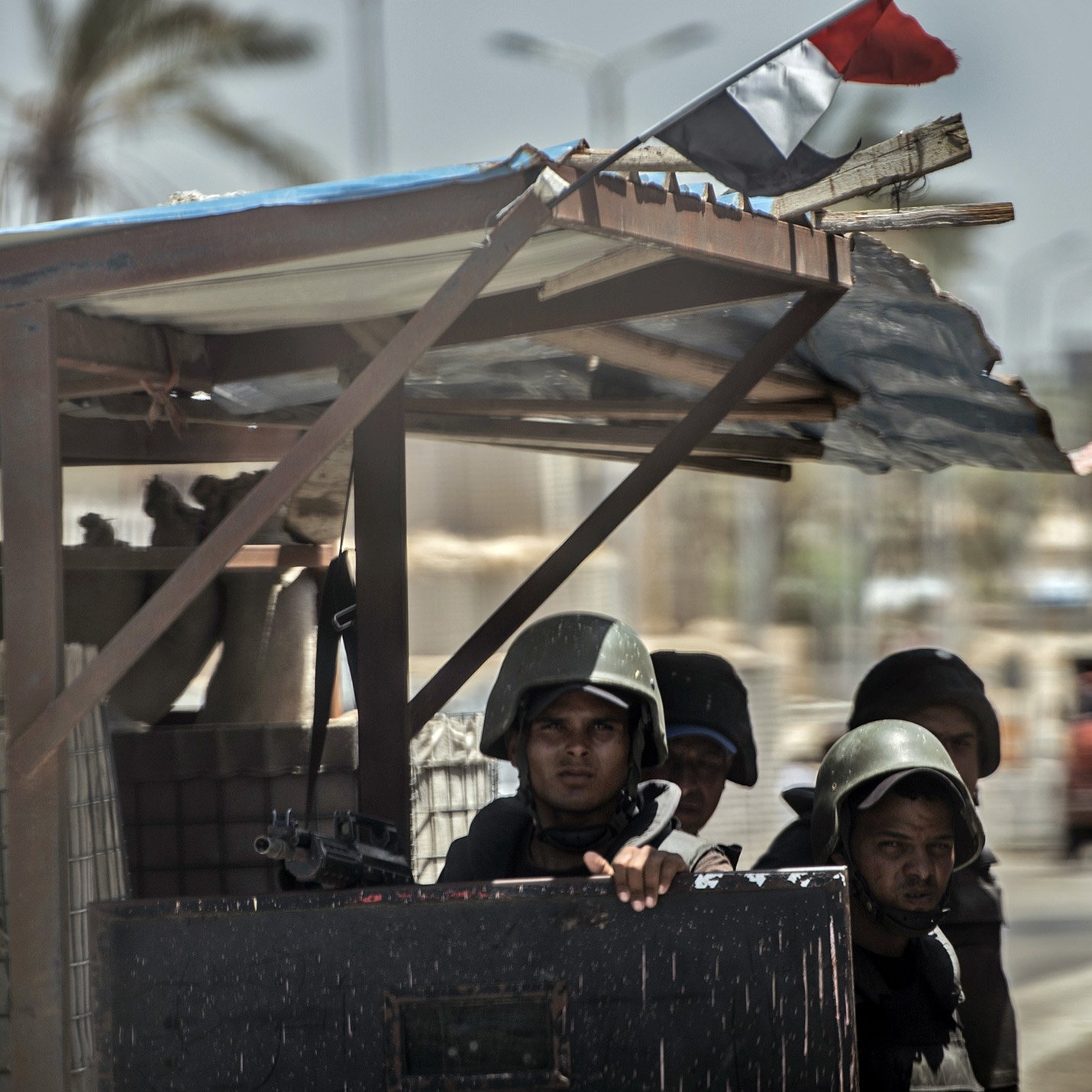The image depicts a group of soldiers stationed at a military outpost. They are wearing helmets and carrying weapons, suggesting they are on alert or guarding a specific area. The backdrop includes palm trees and some vehicles, indicating an urban environment. A flag can be seen on top of the structure, adding to the military context of the scene. The overall atmosphere appears tense and focused on security.