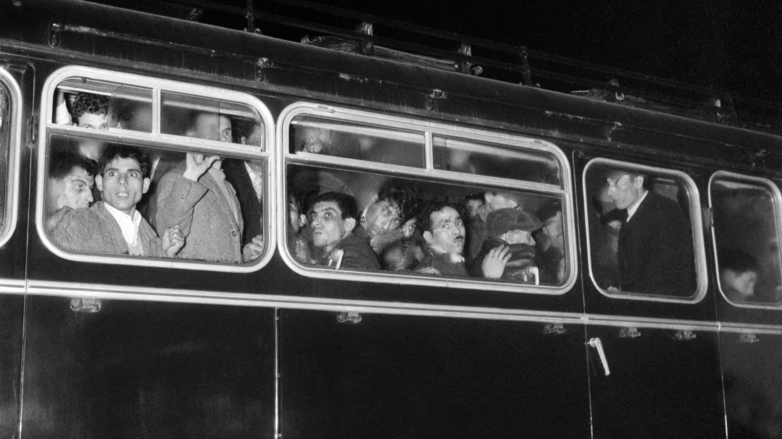 L'image montre un bus noir en soirée, dont les fenêtres sont remplies de passagers. On peut voir plusieurs visages, certains semblent surpris ou curieux, tandis que d'autres affichent des expressions neutres. L'éclairage est faible, accentuant l'atmosphère nocturne de la scène. Le bus a l'air ancien, avec une carrosserie en métal et des détails rétro.
