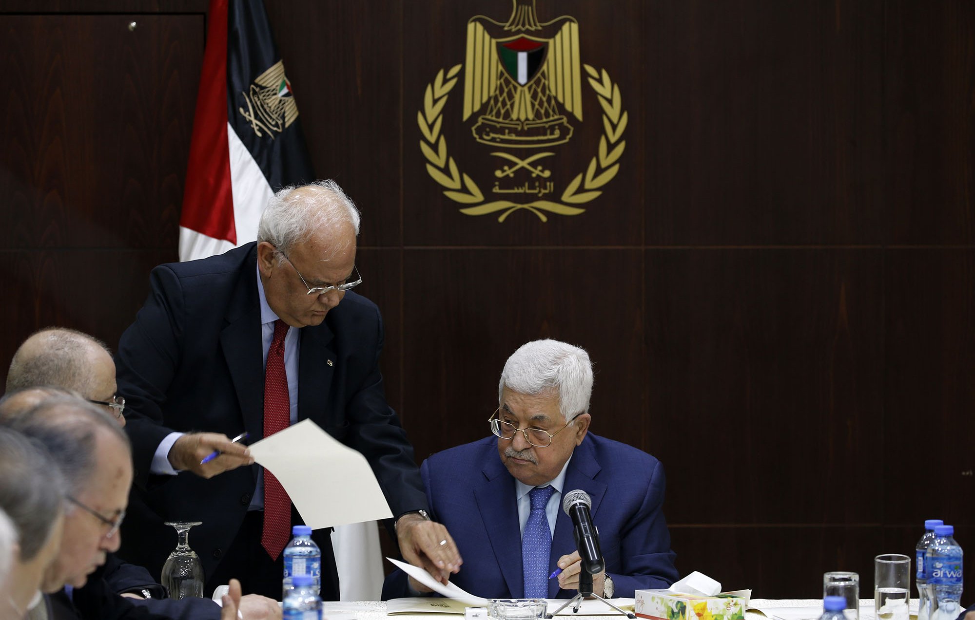 L'image montre une réunion, probablement politique, avec plusieurs hommes assis à une table. Au premier plan, un homme, probablement un leader ou un officiel, est assis regardant des documents. Un autre homme, debout à ses côtés, semble lui présenter des papiers ou des informations. En arrière-plan, on peut voir un drapeau, ainsi qu'un emblème qui semble représenter une organisation ou un gouvernement. L'ambiance générale de la scène est sérieuse et attentive, suggérant une discussion importante.