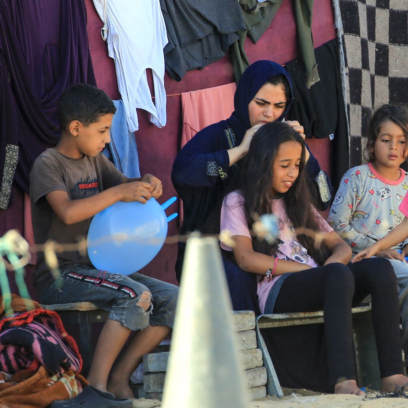 تصویر گروهی از کودکان و یک زن را نشان می‌دهد که در یک محیط غیررسمی نشسته‌اند. زن در حال بافتن یا شانه کردن موی دختری است که روی صندلی نشسته است. سایر کودکان دور آنها در حال بازی یا گفتگو هستند. پس‌زمینه تصویر به وضوح نشان‌دهنده محیطی ساده و روستایی است، با لباس‌های آویخته به دیوار و برخی وسایل دیگر در اطراف. جو تصویر دوستانه و صمیمی به نظر می‌رسد.