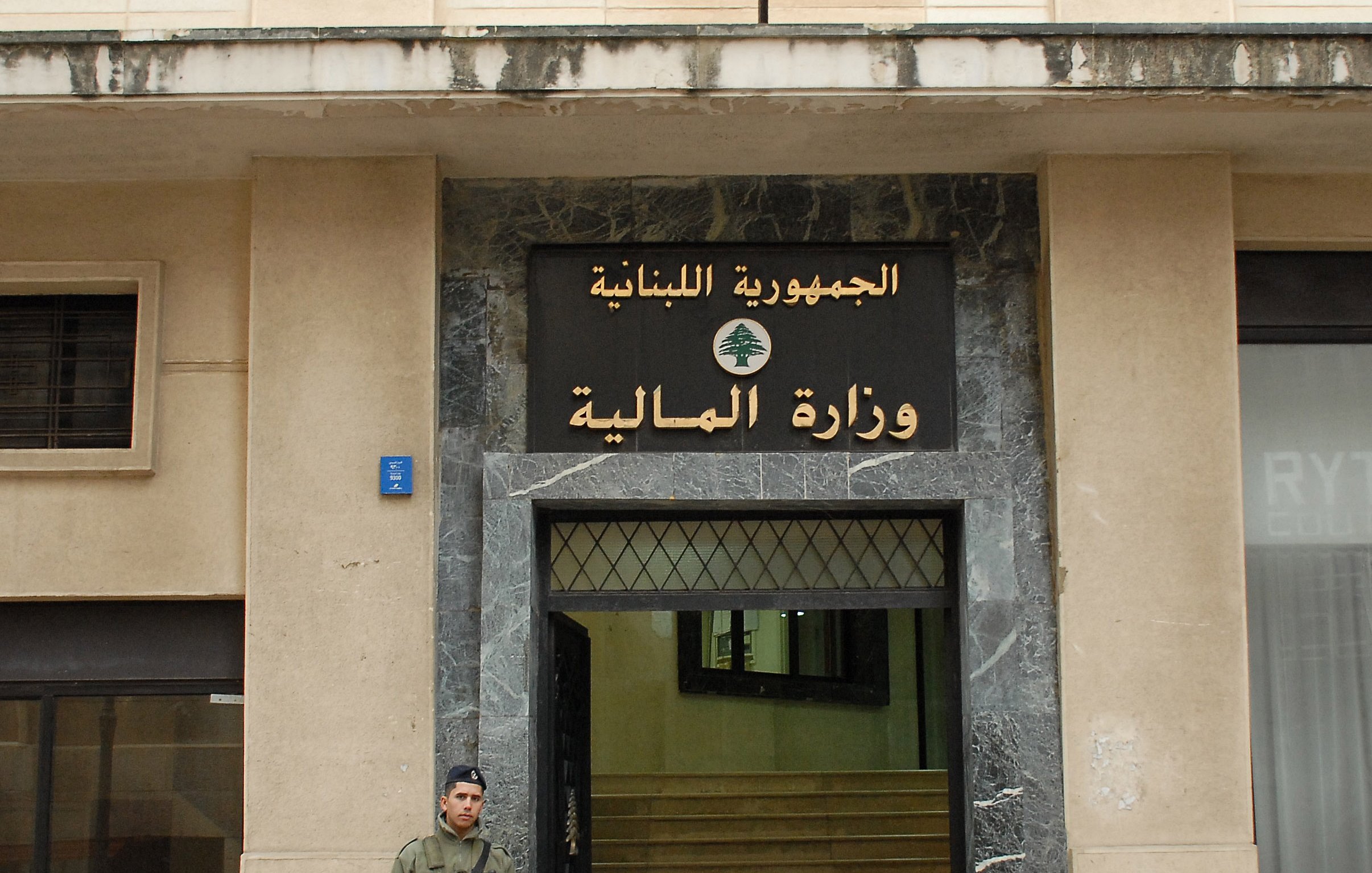 L'image montre un bâtiment avec une façade en pierre, portant une inscription en arabe et en français. On y voit un soldat en uniforme, armé, qui se tient devant l'entrée du bâtiment. Le soldat semble en position de garde, et à côté de lui, on peut remarquer des éléments architecturaux typiques des bâtiments gouvernementaux. L'inscription indique que l'édifice est le ministère des Finances du Liban.