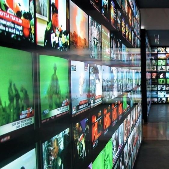 L'image montre une vaste installation composée de nombreux écrans de télévision affichant différentes vidéos et images. Les écrans sont regroupés sur des murs, créant une mosaïque visuelle. Certaines chaînes semblent diffuser des nouvelles, tandis que d'autres présentent des contenus variés. L'éclairage est vif, et l'ensemble donne une impression de dynamisme et de surcharge d'information. L'environnement pourrait évoquer un espace dédié aux médias ou à l'art numérique.