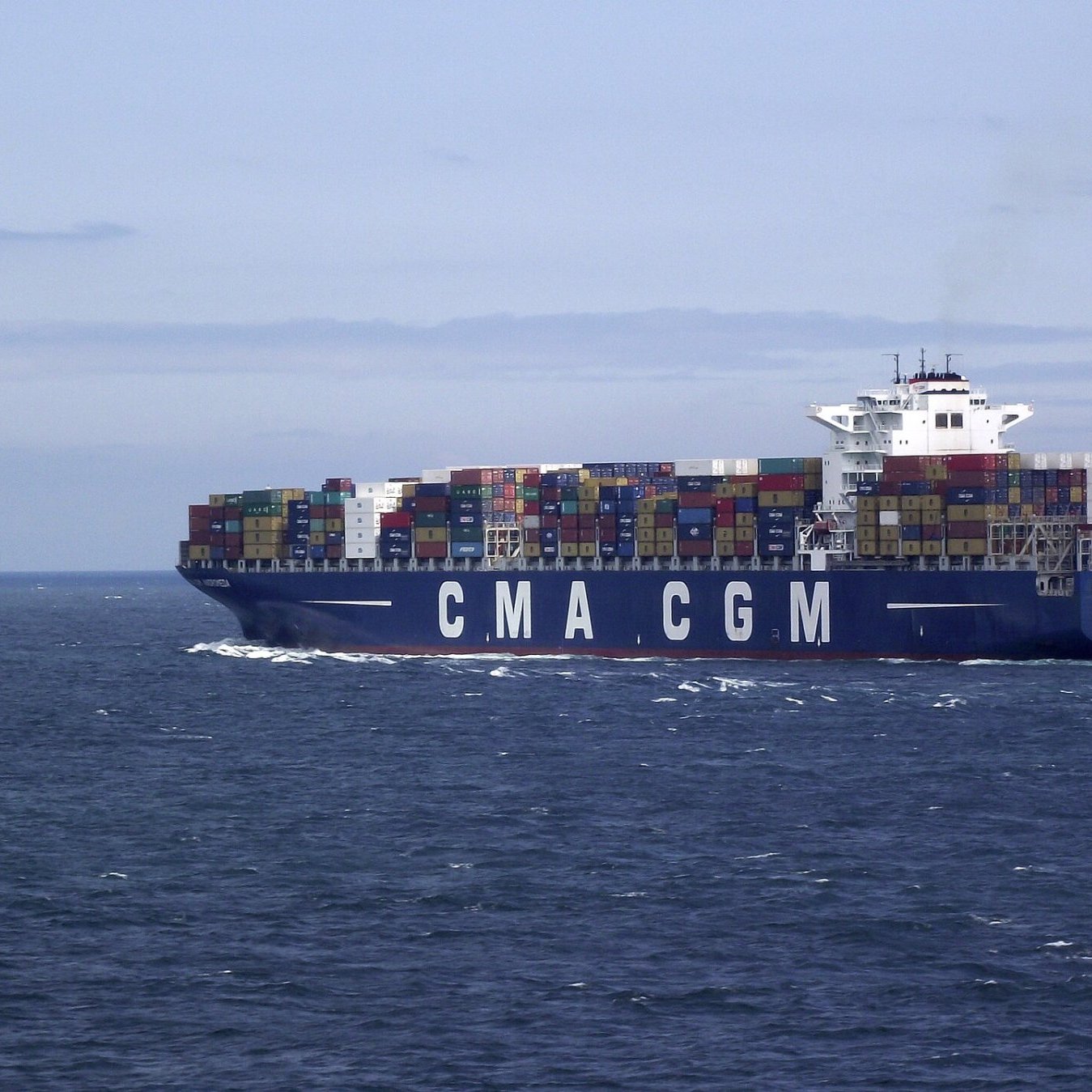 L'image montre un grand porte-conteneurs naviguant sur une mer calme. Le navire, qui porte les lettres "CMA CGM" sur son flanc, est couvert de conteneurs multicolores empilés sur le pont. Le ciel est légèrement nuageux, avec des teintes de bleu, et on peut apercevoir des vagues sur l'eau. L'ensemble dégage une impression de commerce maritime et de transport international.