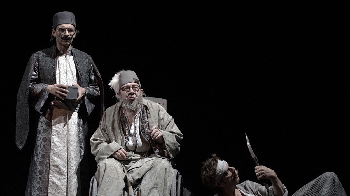 الصورة تظهر مشهداً مسرحياً مع ثلاثة شخصيات. الشخصية الوسطى تجلس على كرسي متحرك، وتبدو في حالة هدوء ورزانة، مرتدية ثياب تقليدية مع عمامة. الشخصيتان الأخريتان تقفان بجانبه، حيث يظهر أحدهما بزي تقليدي أيضاً ويبدو أكثر جدياً، بينما الآخر يظهر بشكل غير مستقر وتمزق في ملابسه، كما يحمل خنجراً. الإضاءة في الخلفية داكنة، مما يبرز الشخصيات في الصورة.