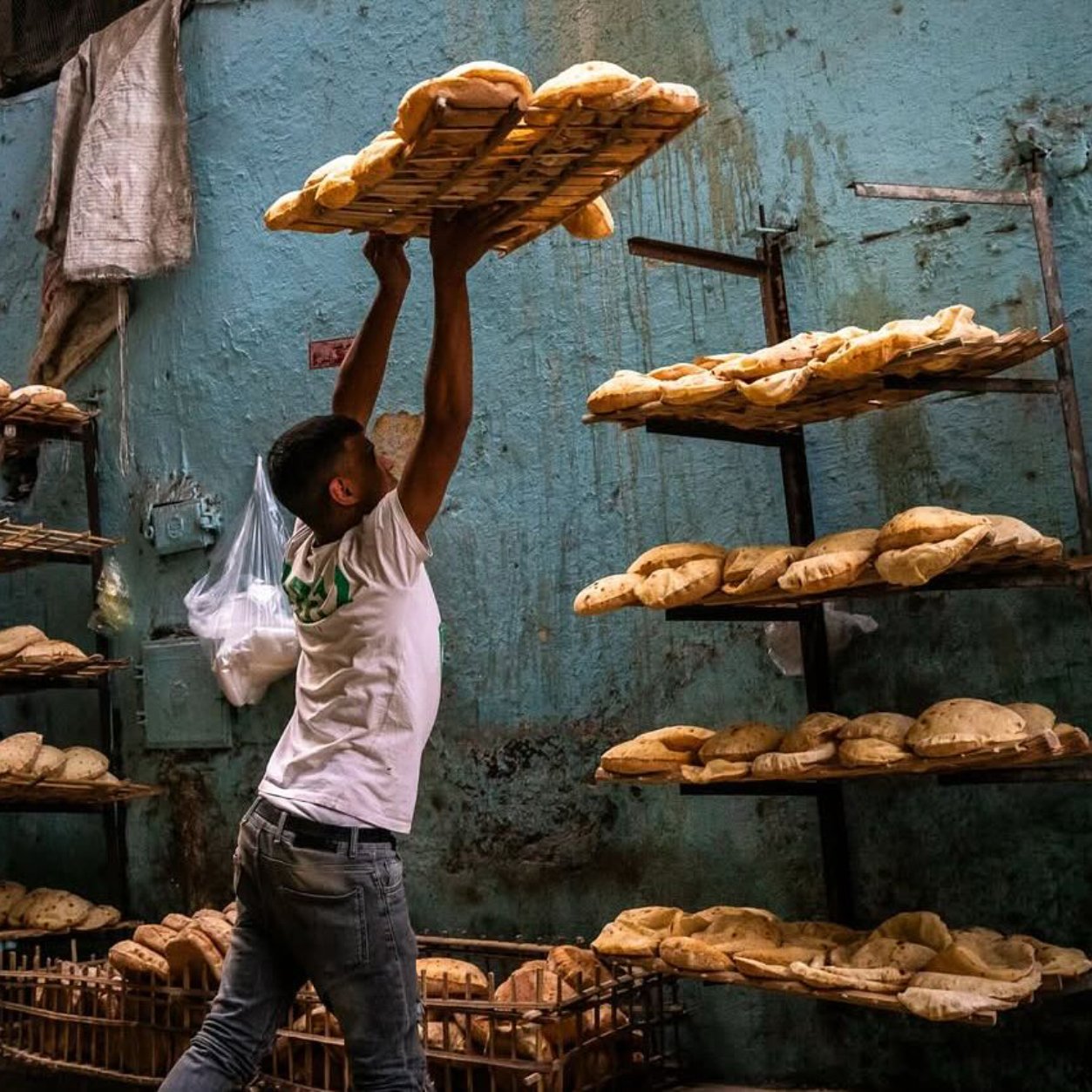 Un boulanger soulève un plateau de pains dans une boulangerie sur un mur bleu.
