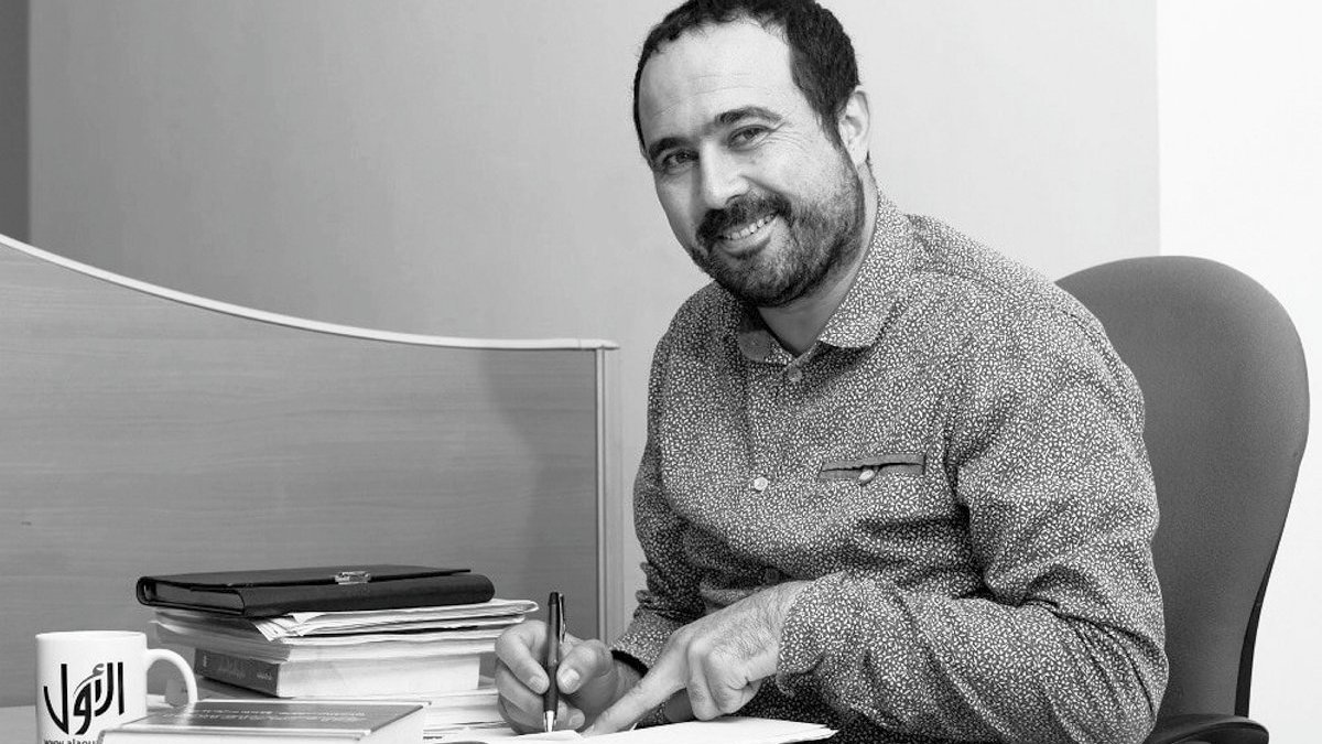 L'image montre un homme assis à un bureau, souriant et tenant un stylo dans une main tout en feuilletant un carnet ou un livre avec l'autre main. Des piles de livres sont présentes sur le bureau, ainsi qu'une tasse à café posée à côté de lui. L'environnement semble être un bureau, avec un fond minimaliste. L'image est en noir et blanc, ce qui lui donne un aspect classique.