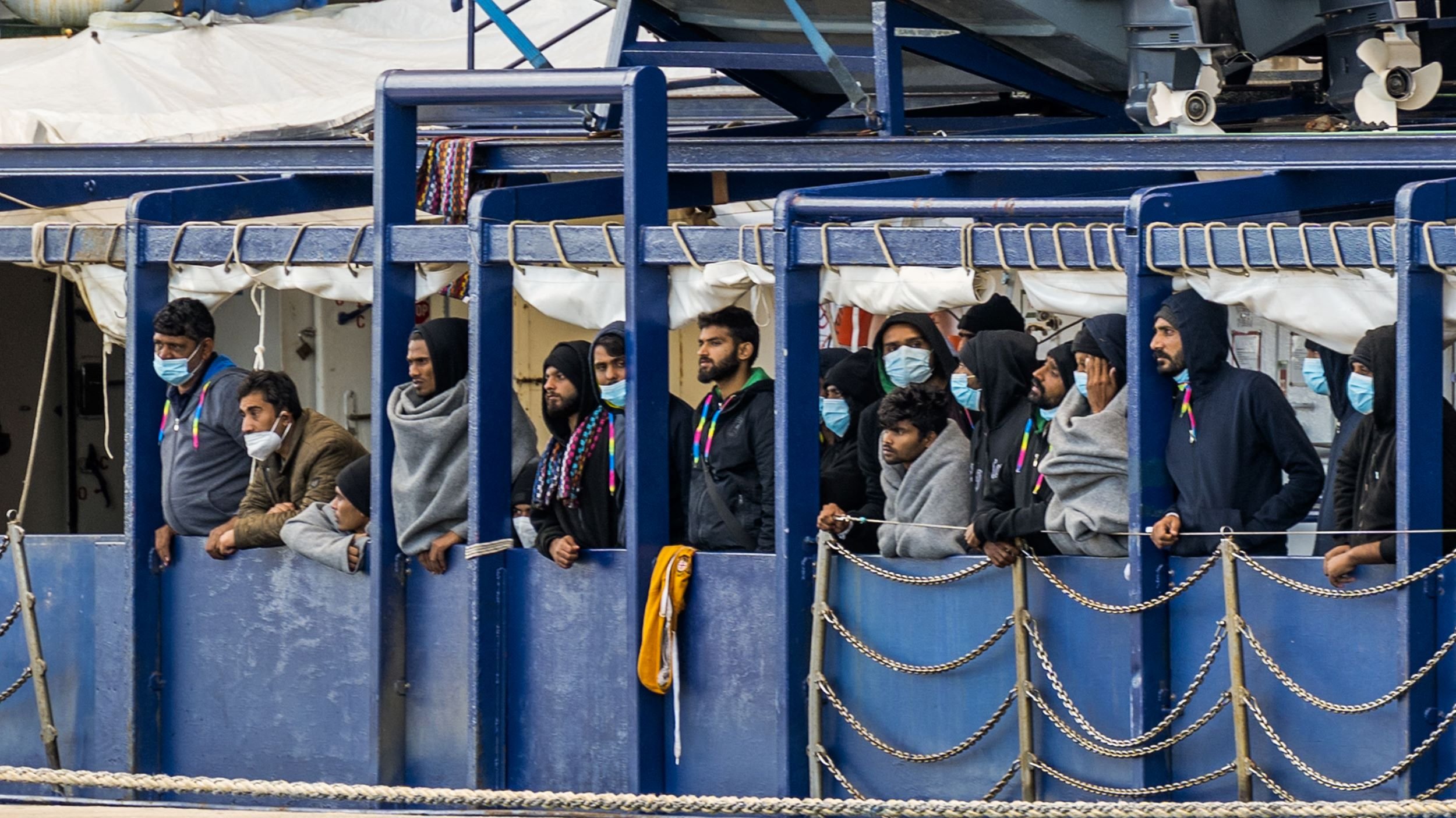 در تصویر، گروهی از افراد در یک محیط بسته مشغول تماشا هستند. آن‌ها ماسک‌های بهداشتی به چهره دارند و برخی از آن‌ها لباس‌های گرم و کلاه بر سر دارند. به نظر می‌رسد که آن‌ها در کنار یک کشتی یا قایق ایستاده‌اند و به چیزی در دوردست نگاه می‌کنند. محیط اطراف شامل سازه‌هایی از فلز و موانع زنجیری است. احساس صمیمیت و توجه در چهره‌هایشان دیده می‌شود.