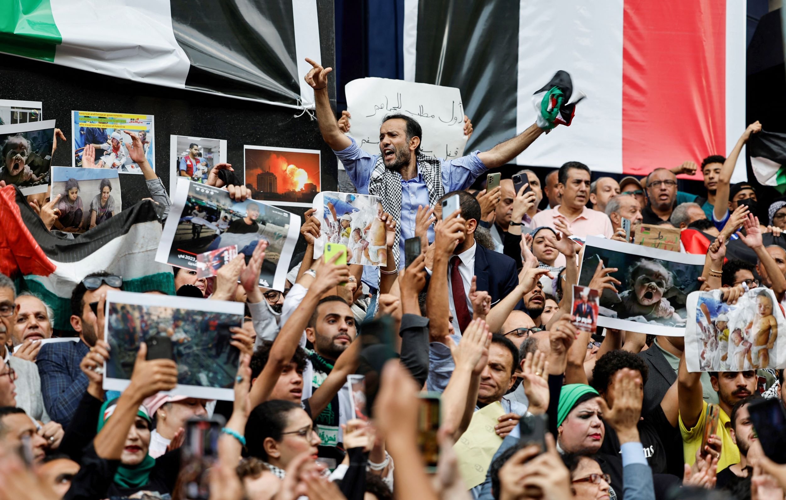 این تصویر یک تجمع بزرگ را نشان می‌دهد که مردم در آن با شور و هیجان حضور دارند. تعدادی از افراد در حال نشان دادن تصاویر و پلاکاردهایی هستند. برخی از معترضان در حال فریاد زدن یا ابراز احساسات خود هستند و مشخص است که موضوعی برای آنها اهمیت دارد. در پس‌زمینه، پرچم‌ها و تصاویری از وقایع یا شخصیت‌های مهم ممکن است مشاهده شود. جو کلی تصویر پر از انرژی و احساسات قوی است.