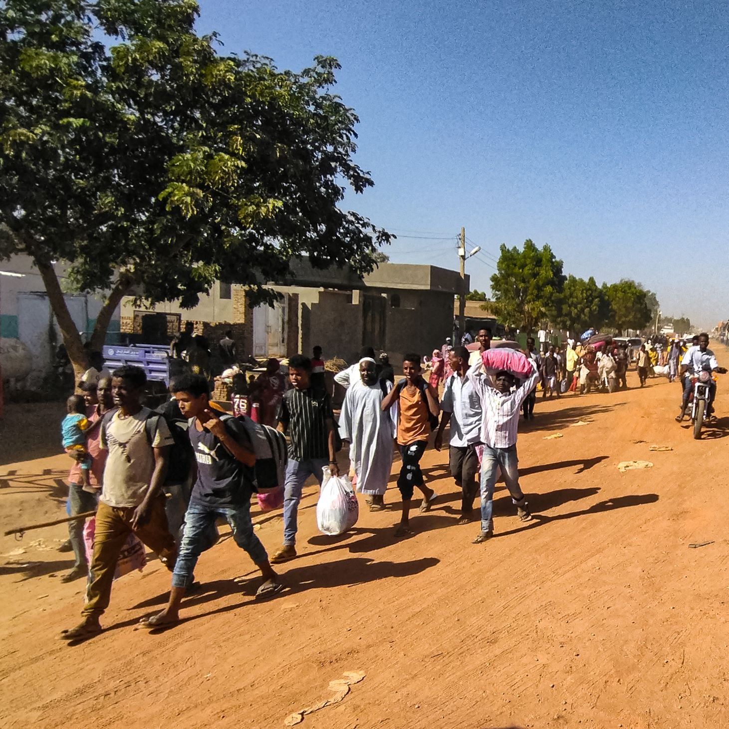 در این تصویر، یک خیابان شنی و پرجنب‌وجوش دیده می‌شود. در سمت چپ، تعدادی درخت و ساختمان‌های ساده قرار دارند. گروهی از مردم در حال پیاده‌روی هستند و برخی از آن‌ها کیسه‌هایی به دوش دارند. در سمت راست، چندین خودرو و موتورسیکلت در حال تردد هستند. فضای عمومی پر از فعالیت و زندگی به نظر می‌رسد.