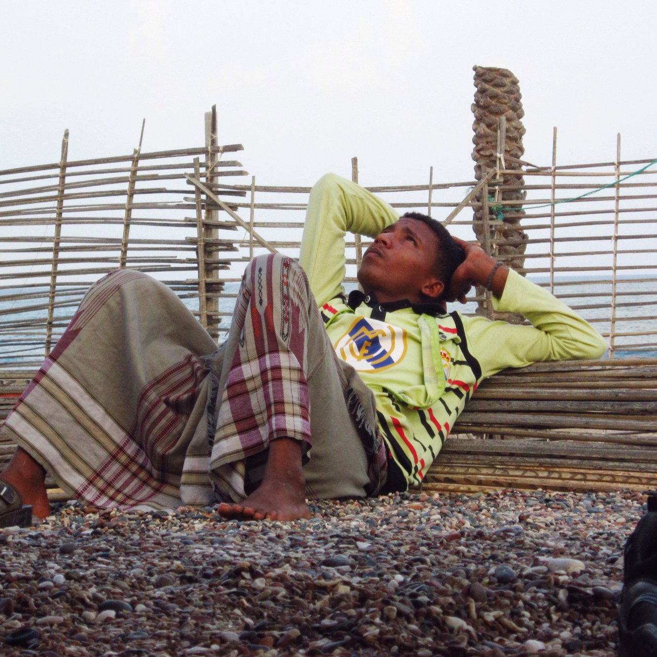 Un garçon allongé, pensif, contre une barrière en bois, sur une plage de gravier.