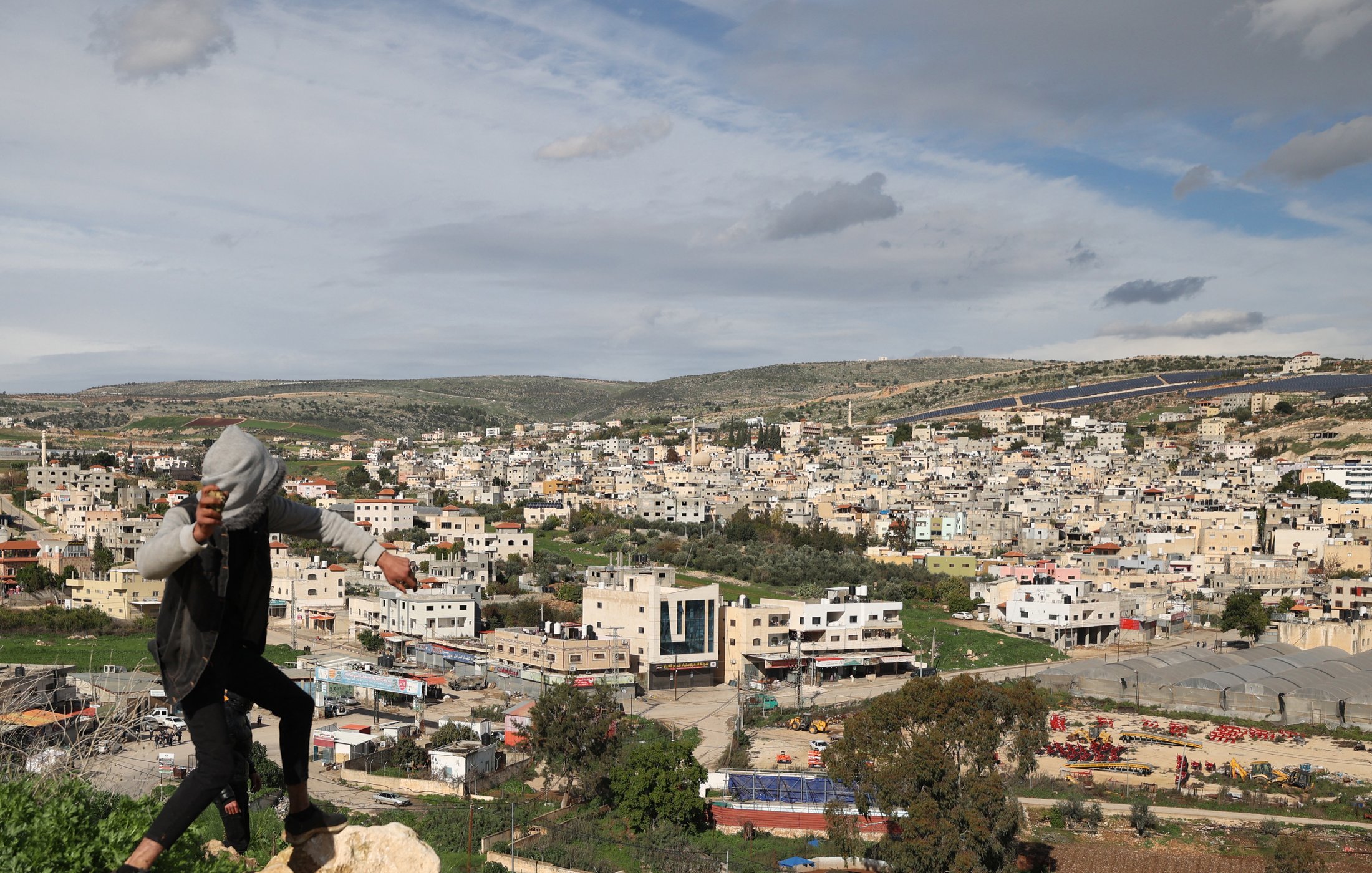 Nell'immagine si vede un giovane che si sta arrampicando su una roccia, con una città sullo sfondo. La città è composta da edifici di diversi piani, molti dei quali di colore chiaro. L'area è circondata da colline verdi e il cielo è parzialmente nuvoloso, suggerendo un clima variabile. Si possono notare anche alcune strutture agricole e aree di terreno aperto in primo piano.