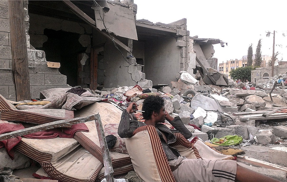 L'image montre une scène de destruction, probablement après un conflit ou une catastrophe naturelle. Au premier plan, un homme assis sur des matelas usés semble perdu dans ses pensées, entouré de débris. À l'arrière-plan, des bâtiments partiellement effondrés et des gravats jonchent le sol, témoignant des conséquences dévastatrices de l'événement. L'atmosphère est sombre et tragique, avec une sensation de désolation.
