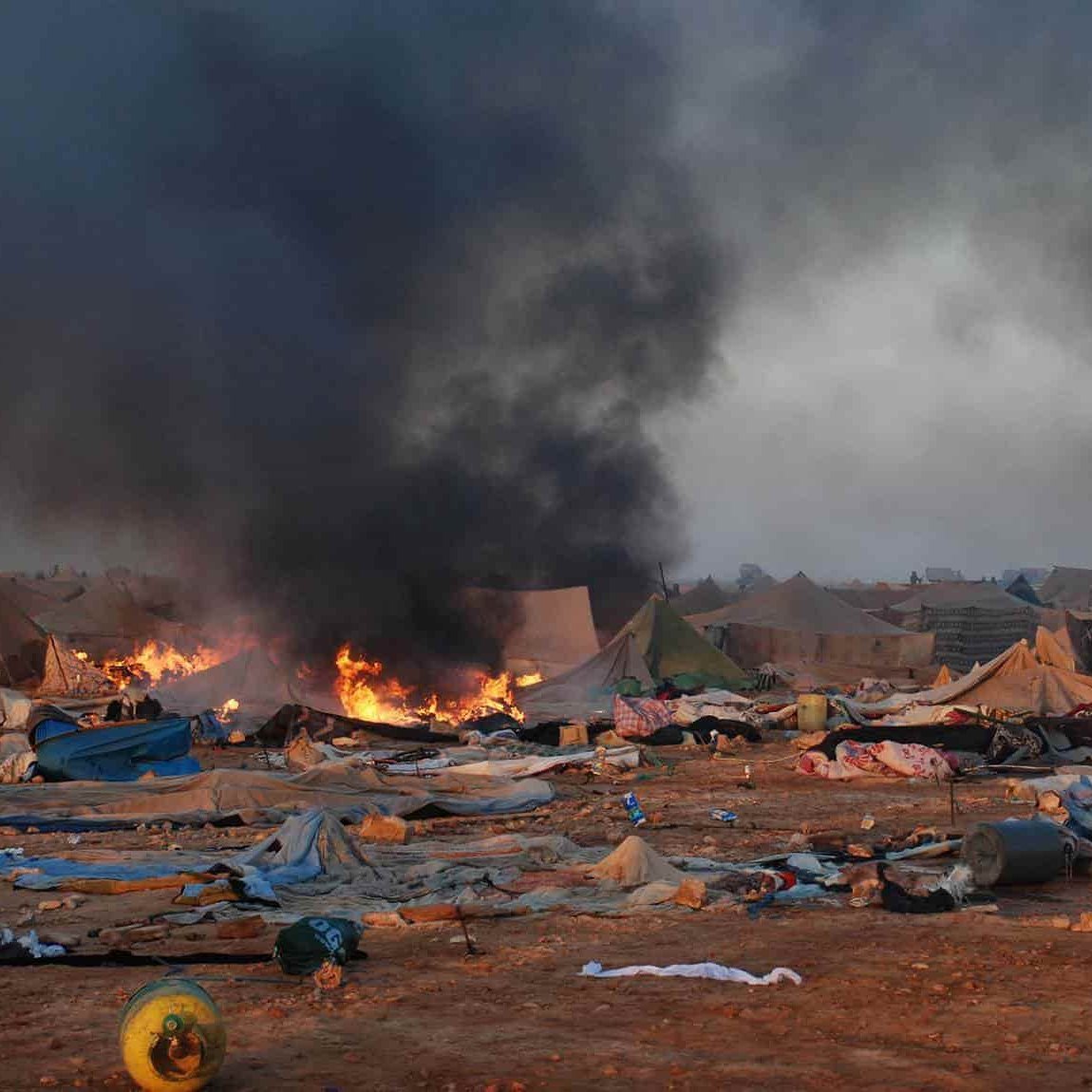 L'image montre un camp de tentes dans un environnement chaotique. On peut voir de la fumée noire s'élever dans le ciel, accompagnée de flammes, indiquant un incendie. Le sol est encombré de débris et de couvertures abandonnées, ce qui suggère une situation difficile. En arrière-plan, des personnes sont visibles, probablement des secouristes ou des autorités, tentant de gérer la situation. L'atmosphère est sombre et perturbée, reflétant une scène de crise ou de catastrophe.