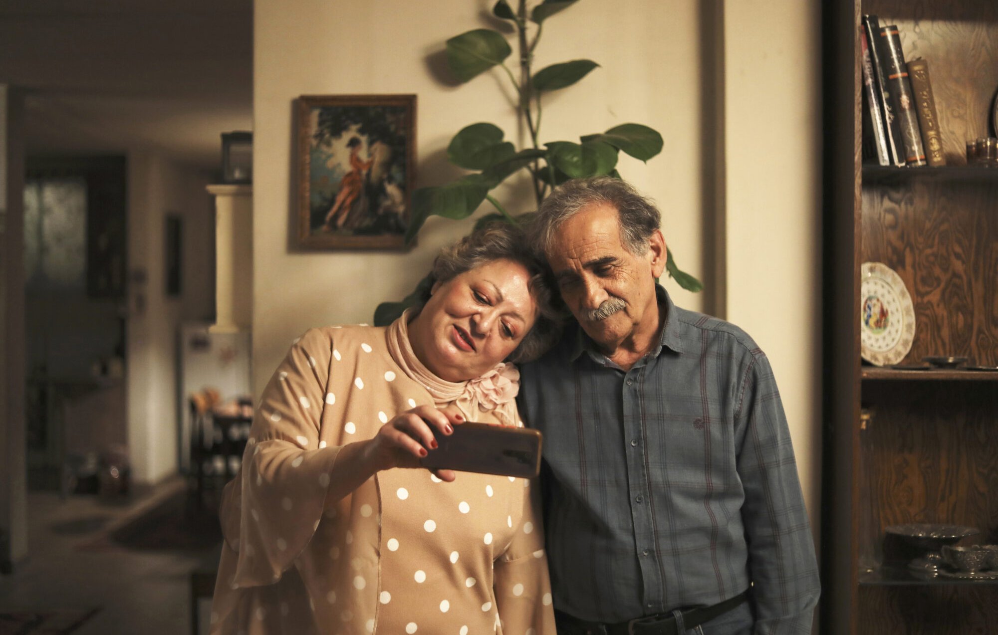 L'image montre un couple âgé, se tenant l'un contre l'autre avec affection. Ils regardent un téléphone ensemble, semblant apprécier ce qu'ils voient. La femme porte une blouse à pois et a un sourire joyeux, tandis que l'homme, en chemise à carreaux, affiche une expression douce. En arrière-plan, on aperçoit un intérieur chaleureux avec des meubles en bois et une plante verte, ce qui crée une ambiance conviviale et intime.