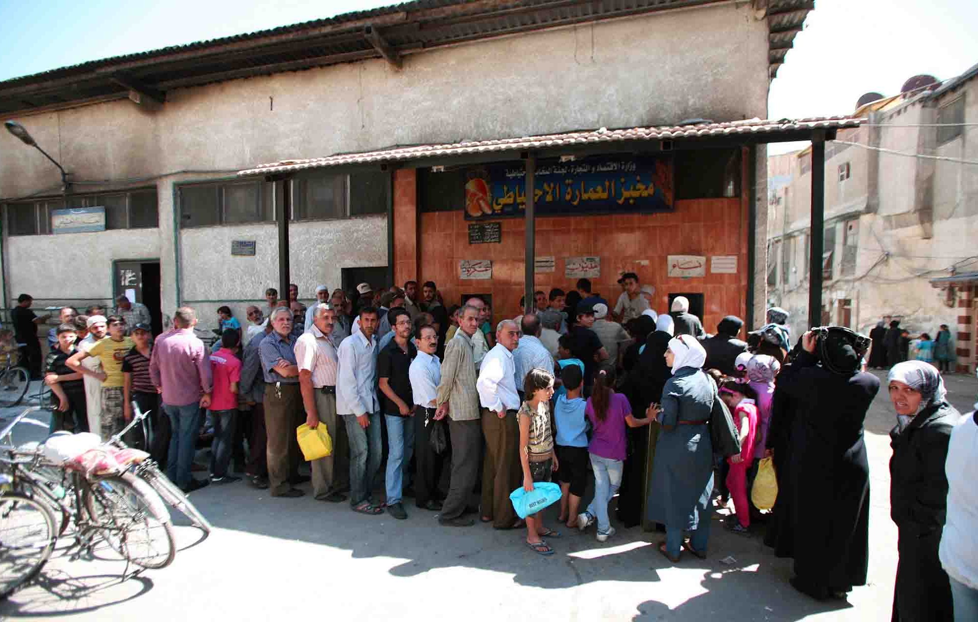 L'image montre une longue file de personnes attendant devant un bâtiment, probablement pour accéder à des services ou des ressources. On peut voir des hommes et des femmes de différentes générations, certains portant des sacs. L'atmosphère semble animée, et le bâtiment est simple, avec une devanture marquée par une enseigne. Des vélos sont stationnés à proximité, et l'environnement urbain est visible en arrière-plan.
