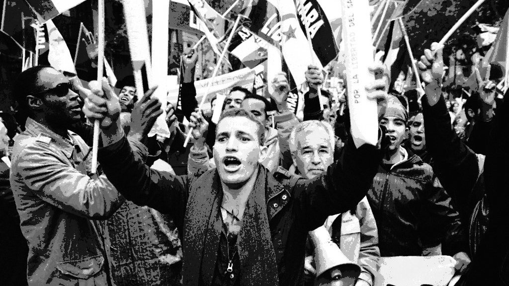 L'image montre une foule en pleine manifestation. On peut voir des personnes brandissant des drapeaux et des pancartes, exprimant leurs opinions avec passion. Au premier plan, un jeune homme s'écrie, tandis que d'autres personnes autour de lui semblent également mobilisées et engagées. L'ambiance générale dégage une forte énergie et un sentiment de solidarité parmi les manifestants. Les visages reflètent une gamme d'émotions, allant de la détermination à l'enthousiasme.
