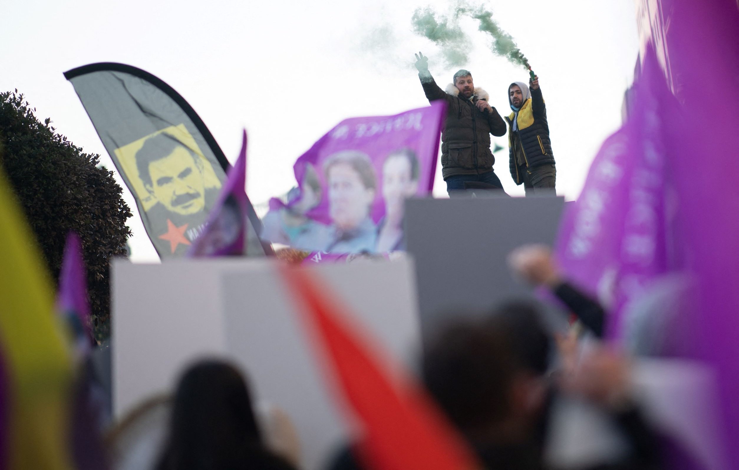 L'image montre une scène de manifestation. Des personnes brandissent des drapeaux et des bannières colorées, principalement en violet et rouge. Au premier plan, on aperçoit deux personnes sur une estrade, l'un d'eux tenant un mégaphone et levant les bras, tandis que l'autre semble encourager la foule. De la fumée verte s'échappe, ajoutant une atmosphère dynamique à l'événement. En arrière-plan, on distingue des portraits, probablement de figures politiques ou symboliques. L'ensemble dégage une ambiance de mobilisation et de passion.