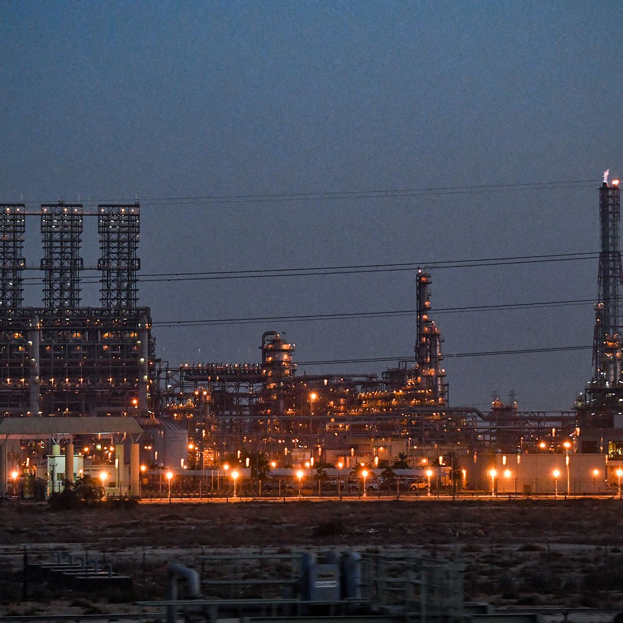 الصورة تُظهر مشهدًا صناعيًا في وقت الغسق، حيث تُضيء الأنوار العديد من المنشآت والمرافق. يمكن رؤية أبراج وكسارات في الخلفية، مع وجود مداخن تدخن في السماء. يبدو أن المنطقة محاطة بإنشاءات كثيرة، مما يشير إلى نشاط صناعي كبير، ويعكس أنوار المصانع انعكاسات الحضارة والتطور التكنولوجي.