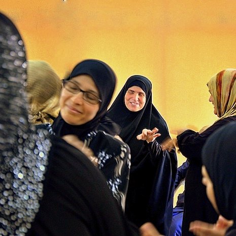 L'image montre un groupe de femmes en discussion ou en interaction. Elles portent des vêtements traditionnels, y compris des hijabs et des abayas. Certaines femmes sourient et semblent engagées dans une conversation joyeuse. L'éclairage dans la pièce donne une ambiance chaleureuse, avec un fond qui semble jaune. L'atmosphère générale est conviviale et animée.