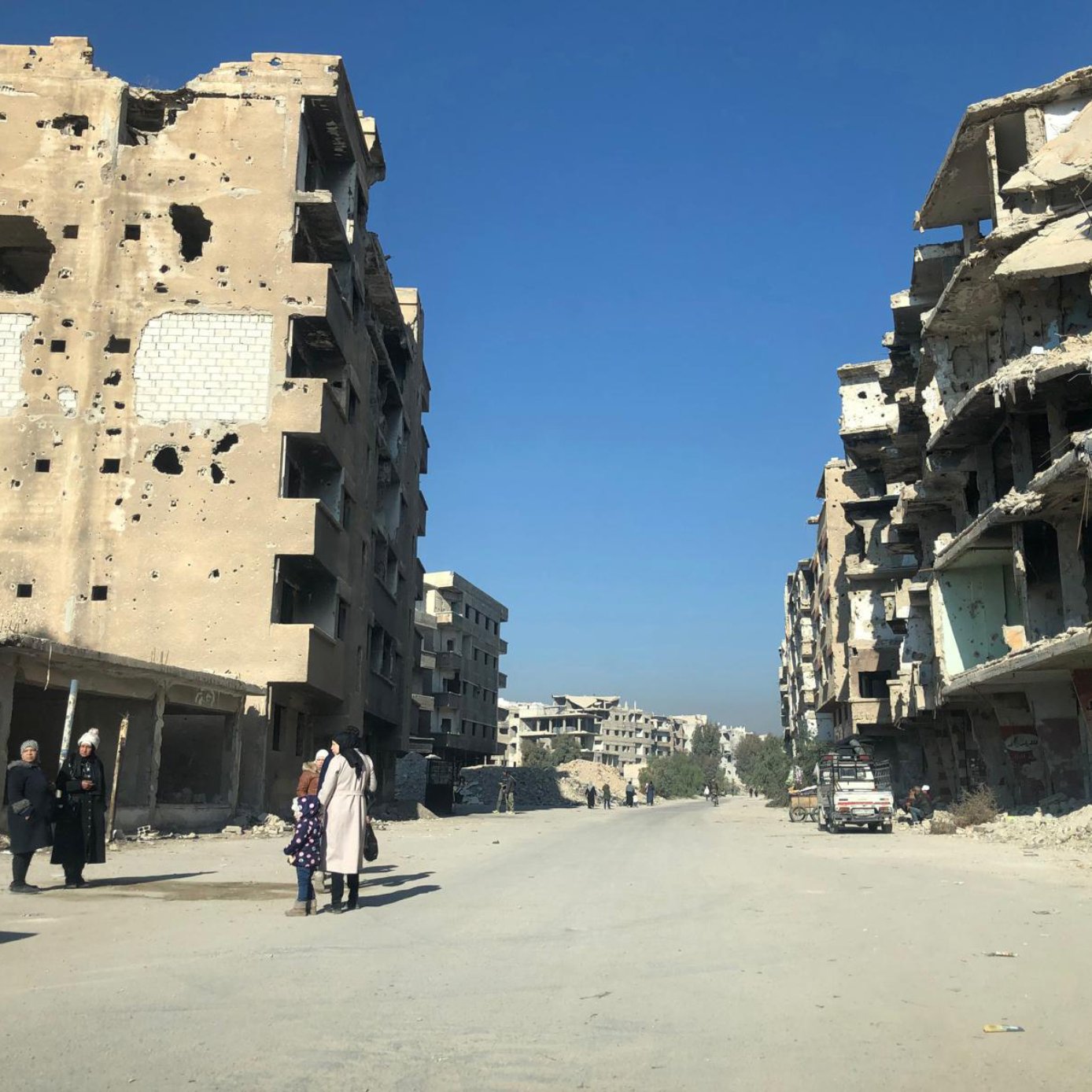L'image montre une rue dévastée par des conflits, avec des bâtiments en ruines de part et d'autre. Les murs des bâtiments sont marqués par des impacts de balles et des débris sont éparpillés sur le sol. Au centre de la rue, quelques personnes marchent, tandis que des voitures abandonnées sont visibles. Le ciel est dégagé, ce qui contraste avec la destruction environnante.