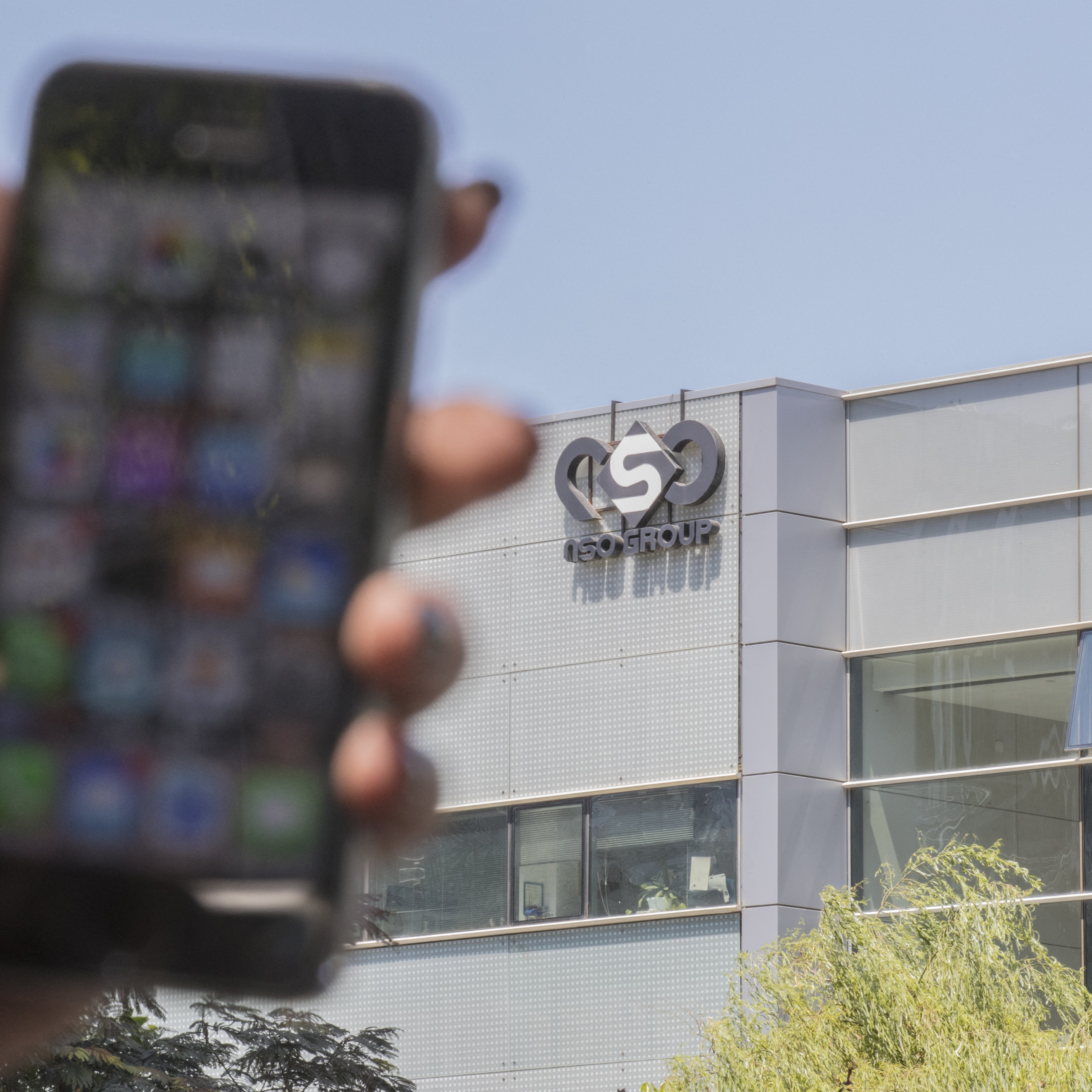 L'image montre un smartphone tenu par une main, flou au premier plan, avec un écran affichant diverses applications. En arrière-plan, on peut voir le bâtiment d'une entreprise, identifié par un logo qui semble être celui d'un groupe nommé "ISN GROUP". Le bâtiment a une façade moderne, probablement en verre. La lumière naturelle éclaire la scène, indiquant qu'il fait jour.