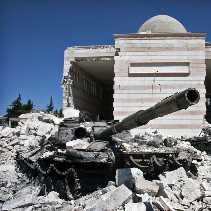 L'image montre un paysage de destruction, probablement lié à un conflit. Au premier plan, on voit des chars endommagés, couverts de débris et de poussière. En arrière-plan, il y a un bâtiment partiellement détruit, dont les murs présentent des marques des destructions. Le ciel est bleu, ce qui contraste avec la scène de désolation au sol. L'ensemble évoque un endroit marqué par la guerre, avec des traces visibles de violence et de désastre.