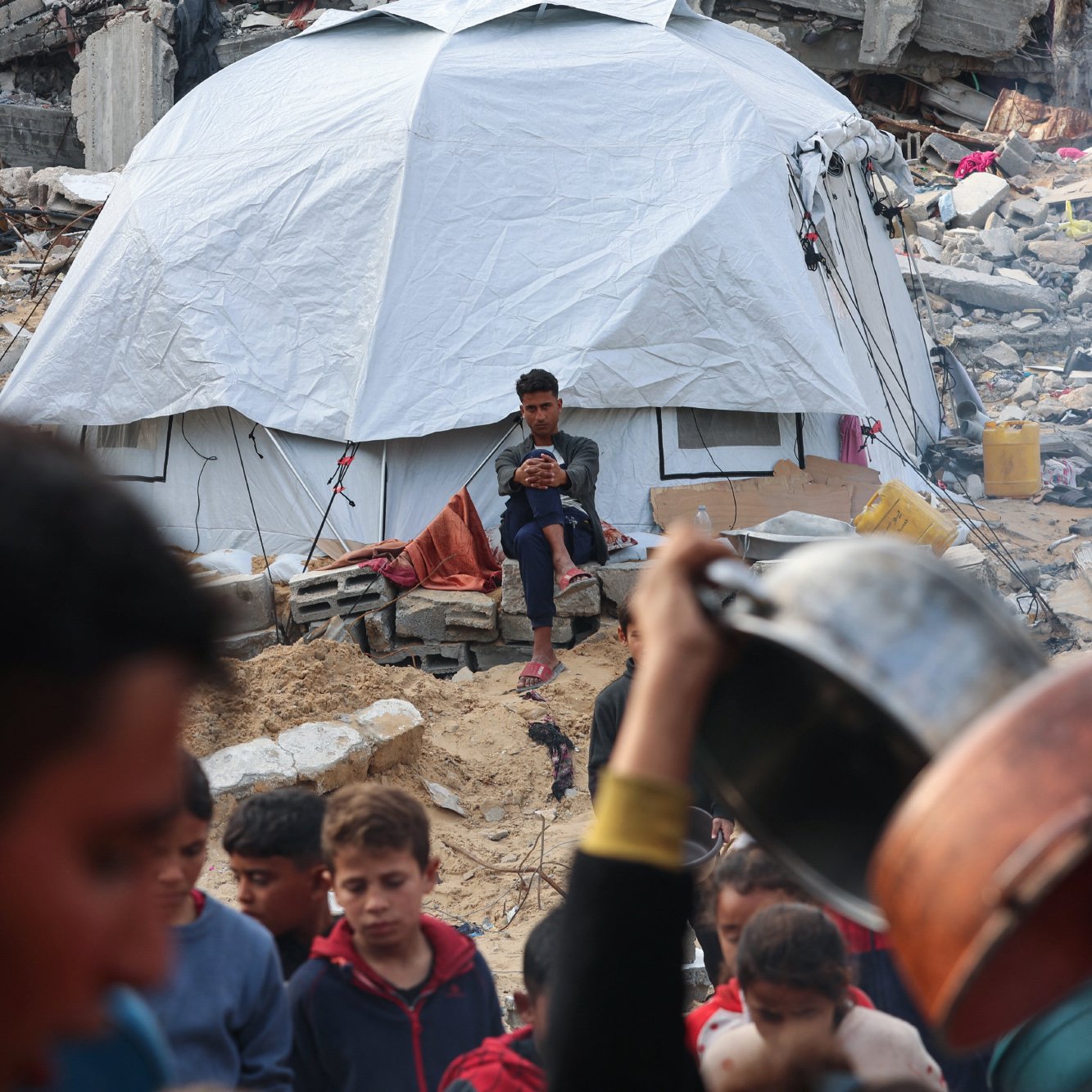L'image montre un camp de réfugiés après un désastre. Au premier plan, un groupe de personnes autour d'une casserole. En arrière-plan, une tente blanche est visible, avec un jeune assis, regardant son téléphone, tandis que des débris de bâtiments sont éparpillés autour.