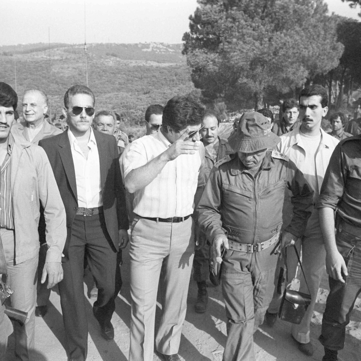 Des hommes en marche, certains en uniforme militaire, sous un ciel dégagé et des arbres.