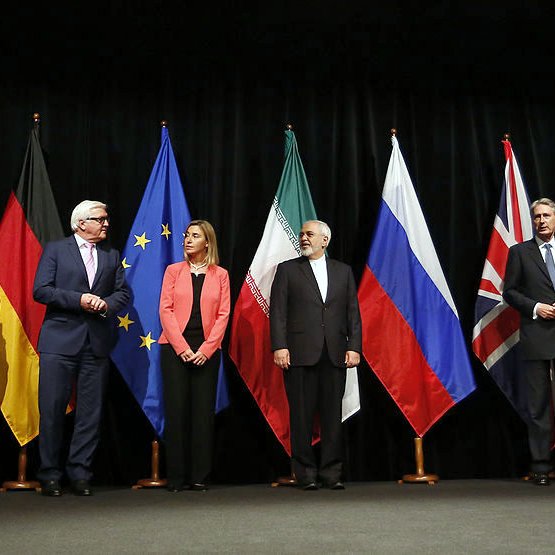 L'image montre une scène officielle avec plusieurs personnes debout en rang, chacune représentant un pays ou une organisation distincte. Derrière eux, il y a des drapeaux de différents pays, notamment la Chine, l'Allemagne, les États-Unis, la Russie et l'Union européenne, entre autres. Les participants semblent engagés dans une discussion ou une négociation, chacun portant un costume formel. L'atmosphère est sérieuse, probablement liée à des pourparlers diplomatiques ou à un sommet international.