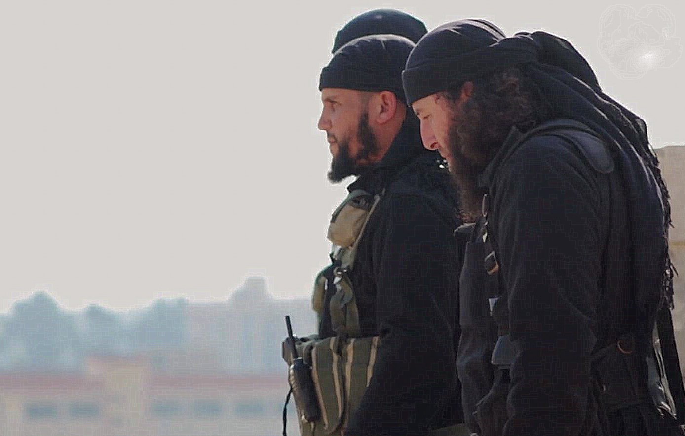 تظهر الصورة مجموعة من الرجال يرتدون ملابس داكنة. يبدو أنهم في وضعية استعداد، حيث يمكن رؤية بعضهم من الجانب. يرتدي البعض منهم شارات أو معدات تكتيكية، مما يدل على أنهم قد يكونون في سياق عمل يتعلق بالأمن أو العمليات العسكرية. خلفهم يظهر مشهد غير واضح، لكن يمكن رؤية بعض المباني في الخلفية. الأجواء عامة تبدو جادة.